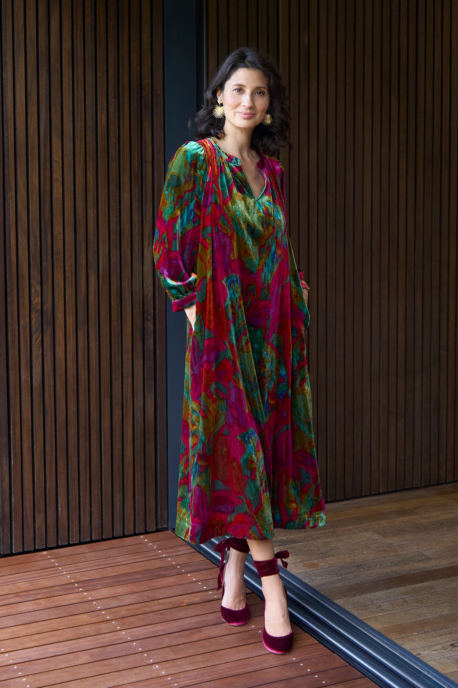 A woman stands indoors against a wooden panel wall, wearing the Velvet Blurred Botanicals Dress with long sleeves and red velvet shoes.