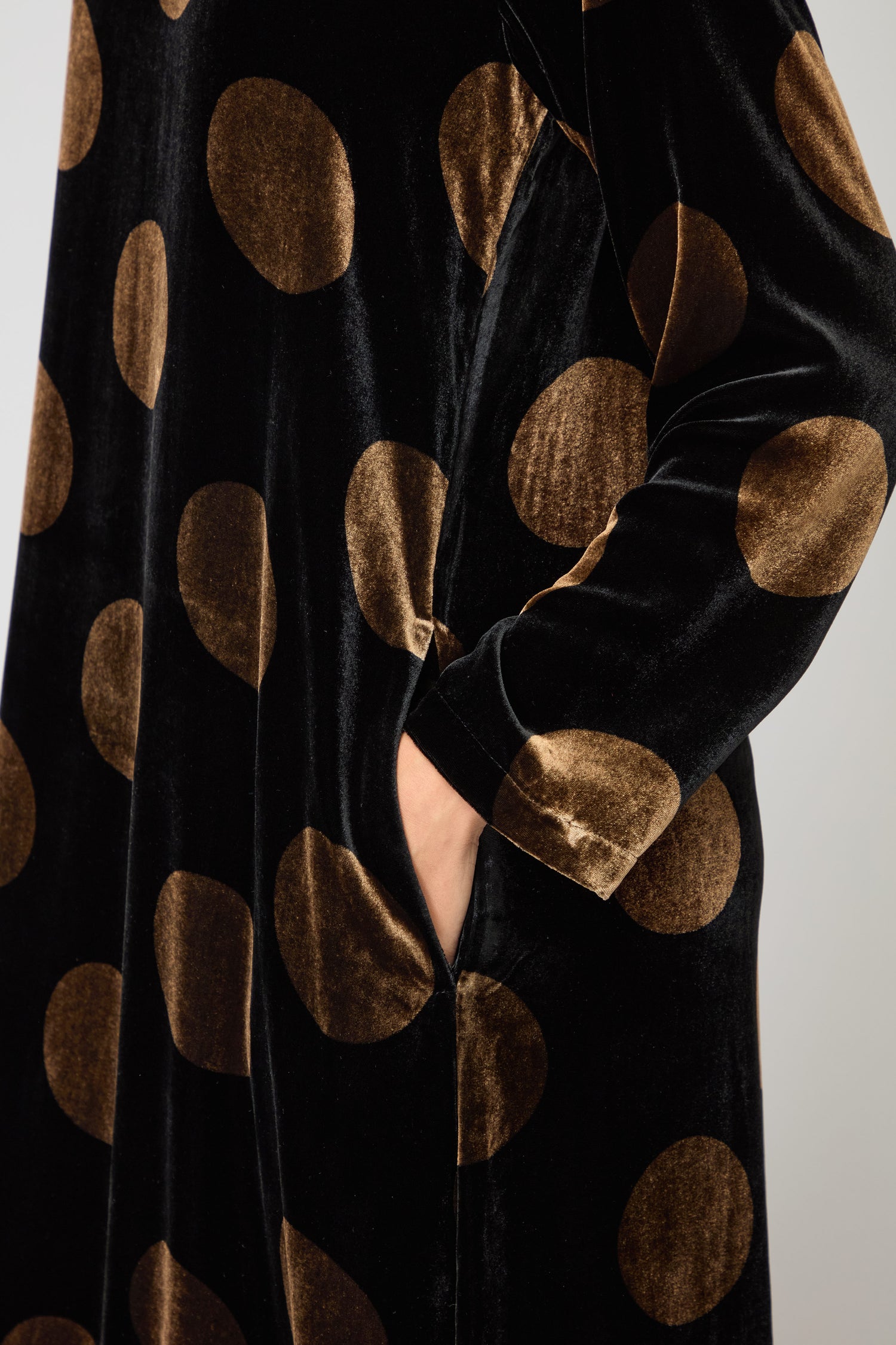 Wearing the Silk Velvet Spot Bubble Dress, which features black velvet fabric with large gold polka dots and subtle botanical motifs, a person stands with their hand in a side pocket.