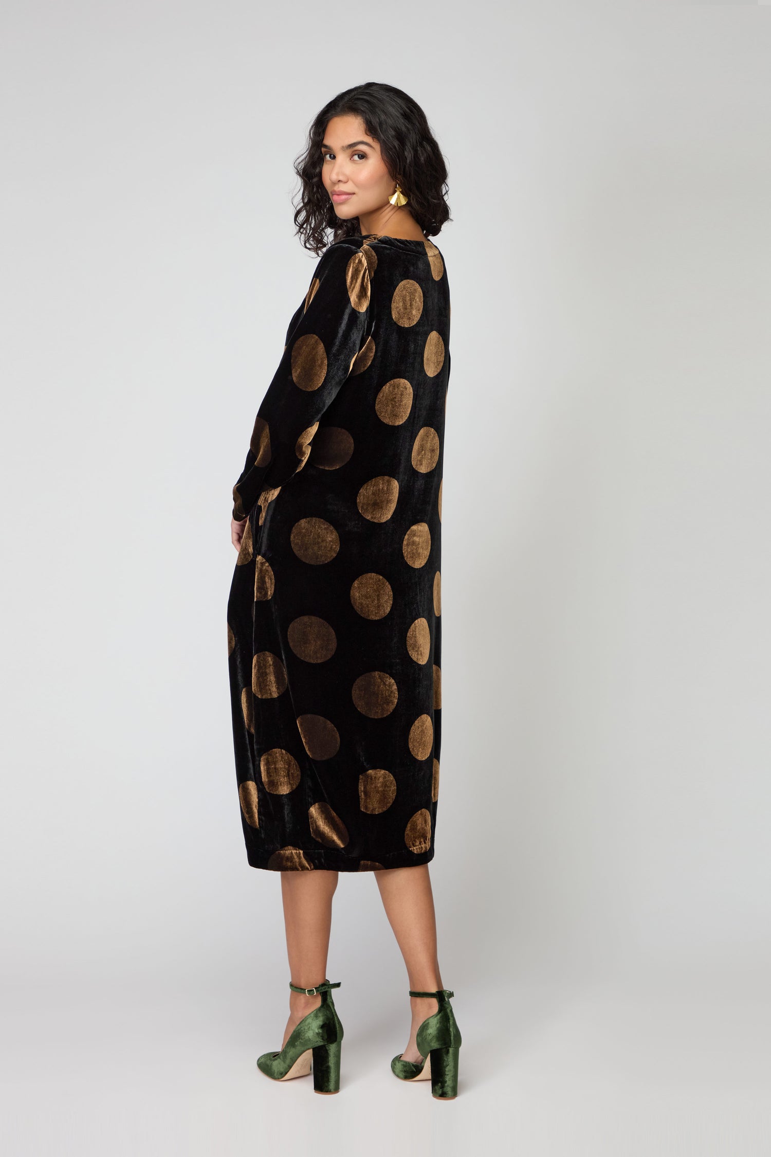 A woman stands turned to the side, wearing the Silk Velvet Spot Bubble Dress with large gold polka dots and green high-heeled shoes, set against a plain light background.