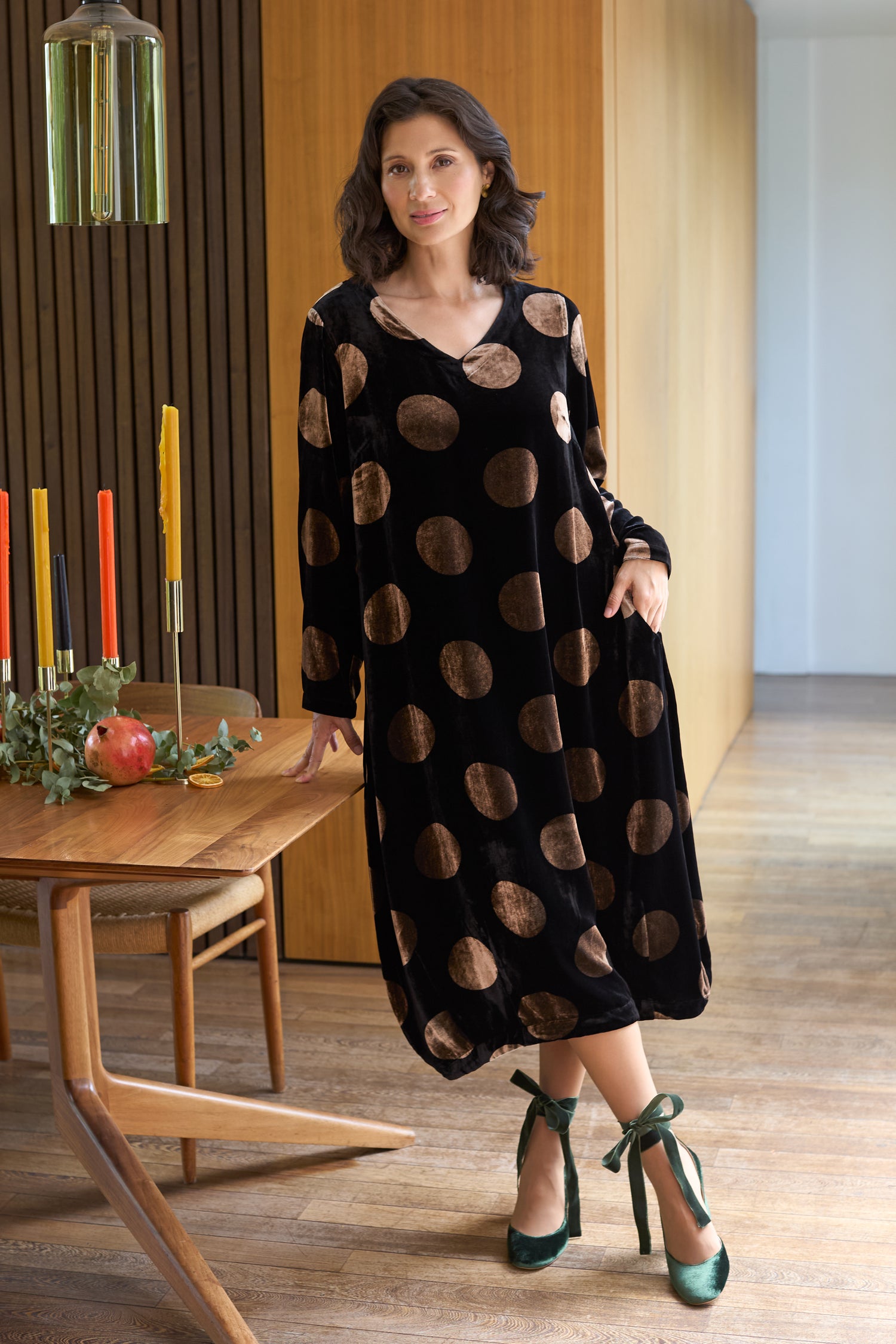 A woman stands indoors wearing the Silk Velvet Spot Bubble Dress, a long black dress with large brown polka dots, paired with green ribboned heels beside a wooden dining table set with candles and a pomegranate.