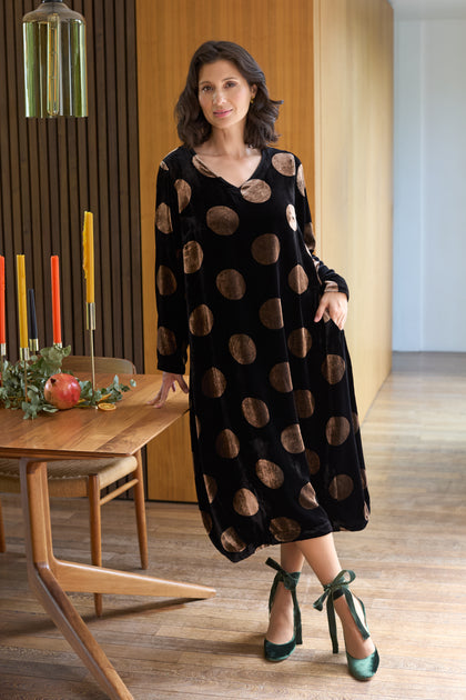 A woman stands indoors wearing the Silk Velvet Spot Bubble Dress, a long black dress with large brown polka dots, paired with green ribboned heels beside a wooden dining table set with candles and a pomegranate.
