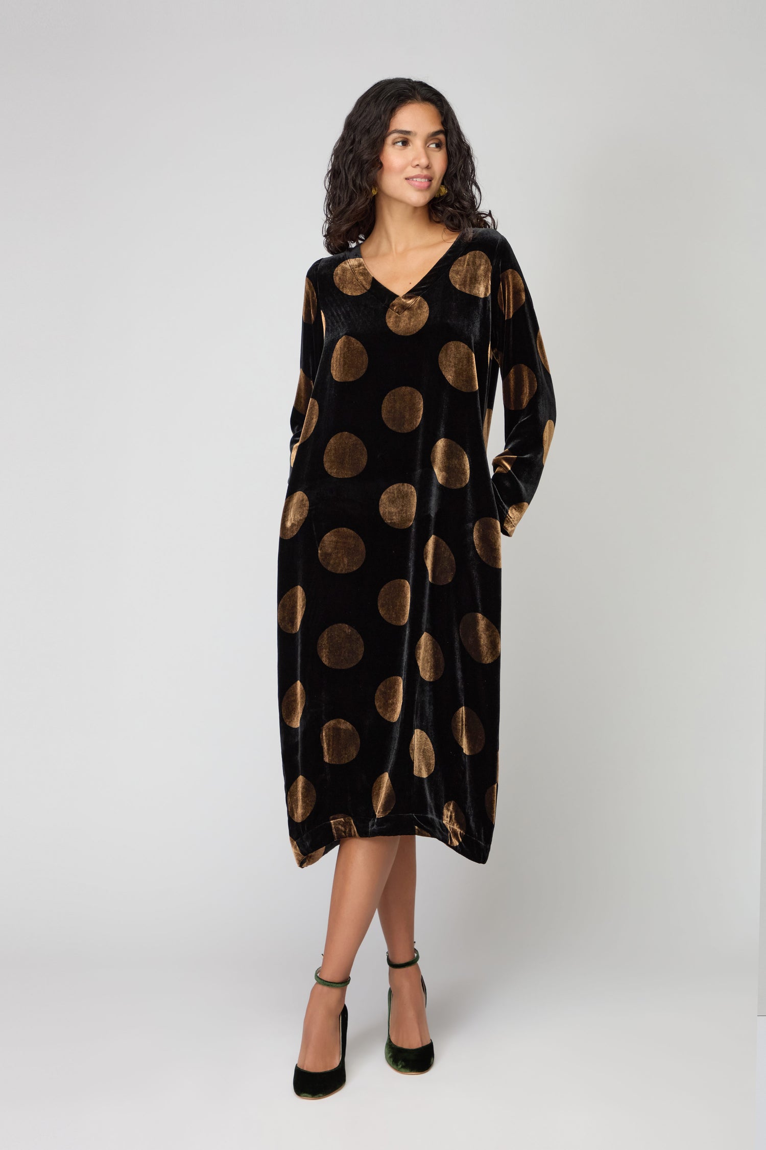 A woman wearing the Silk Velvet Spot Bubble Dress, a loose black velvet dress with large gold dots, long sleeves, and a V-neckline, stands against a plain light background.