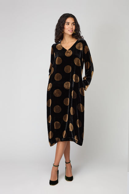 A woman wearing the Silk Velvet Spot Bubble Dress, a loose black velvet dress with large gold dots, long sleeves, and a V-neckline, stands against a plain light background.