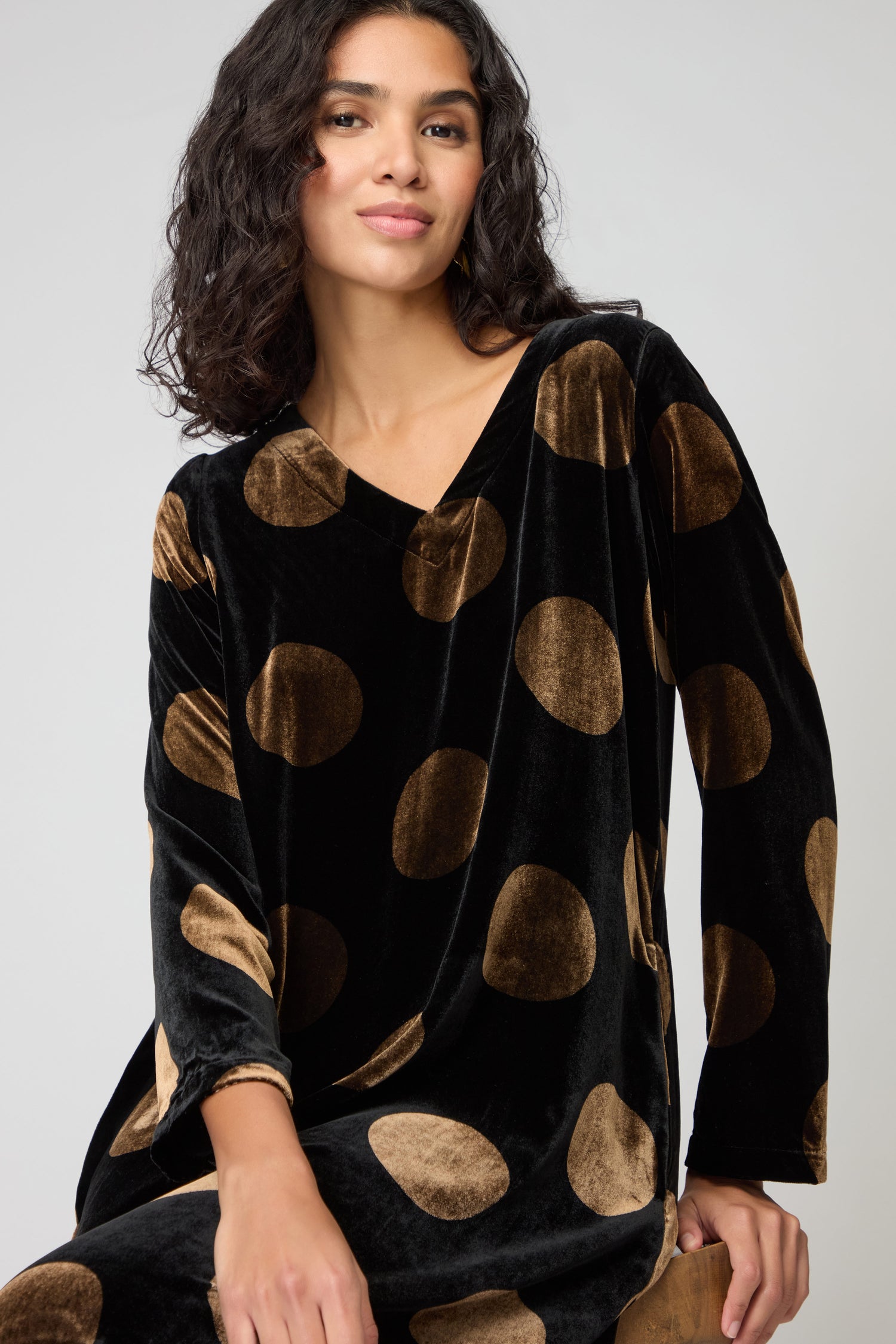 Woman with wavy dark hair wearing the Silk Velvet Spot Bubble Dress, featuring large brown polka dots, seated on a stool against a plain light background.