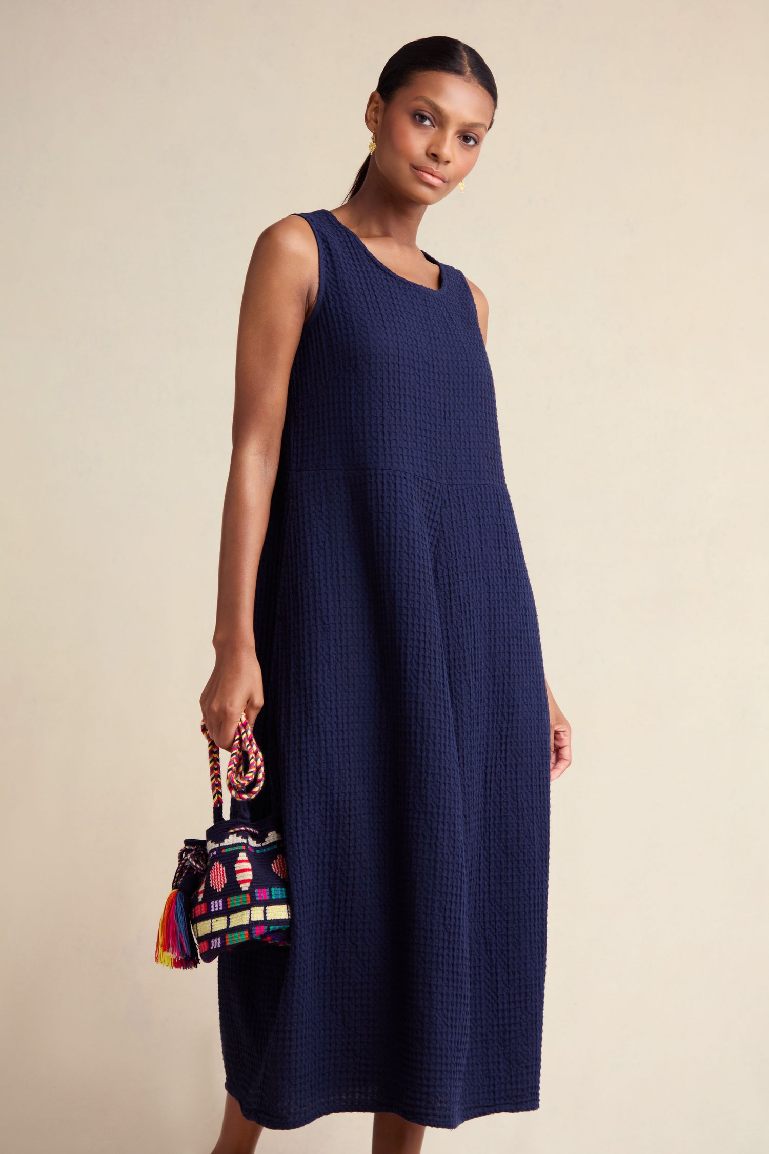 A woman stands against a plain light background wearing the Mini Waffle Cotton Bubble Dress, a sleeveless navy blue textured weave midi, and holds a small colorful embroidered handbag.