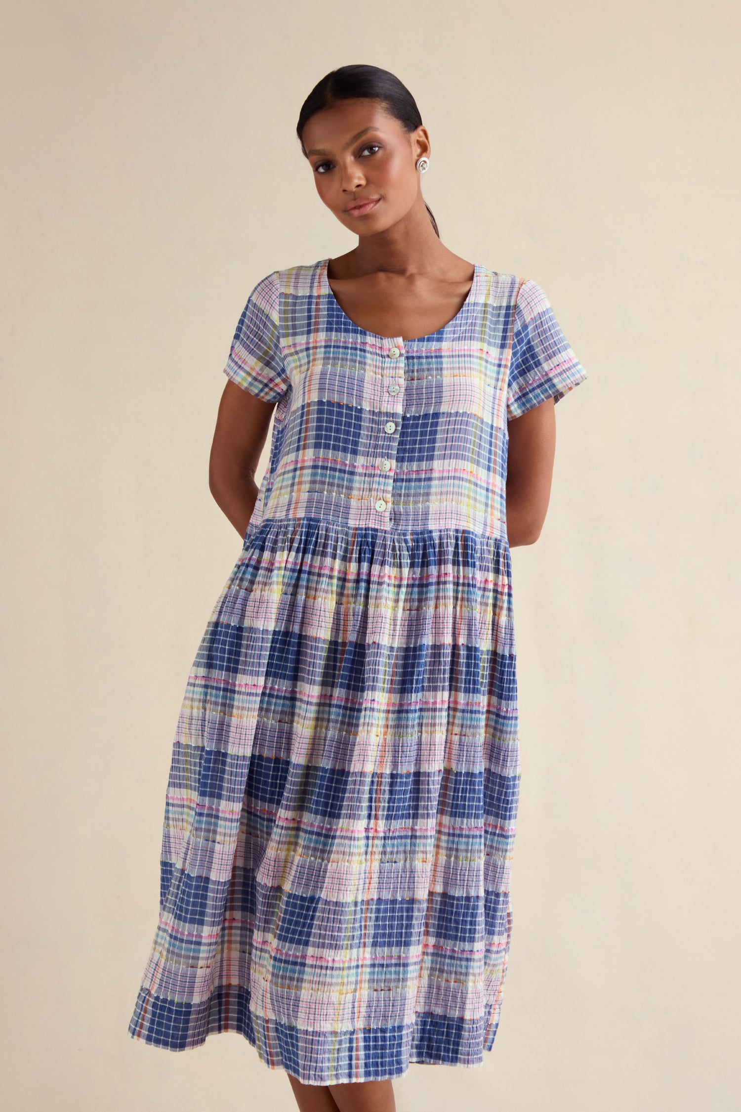 A woman stands in front of a plain, light background wearing the Crinkle Check Jacquard Dress, featuring short sleeves, a button-front, an A-line silhouette, and a gathered skirt.