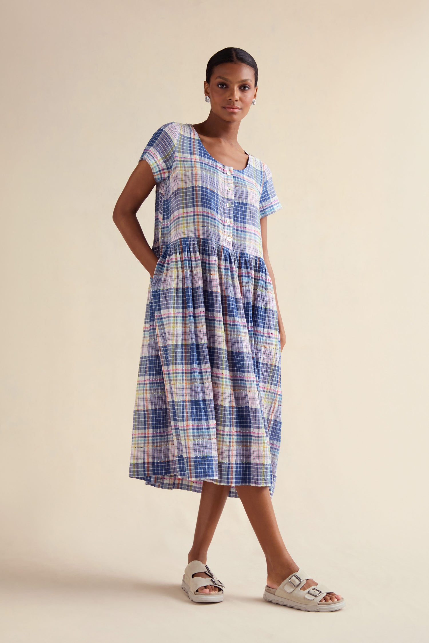 A woman in the Crinkle Check Jacquard Dress, a cotton-rich, short-sleeve, button-front midi with side pockets, and white sandals stands against a light beige background.