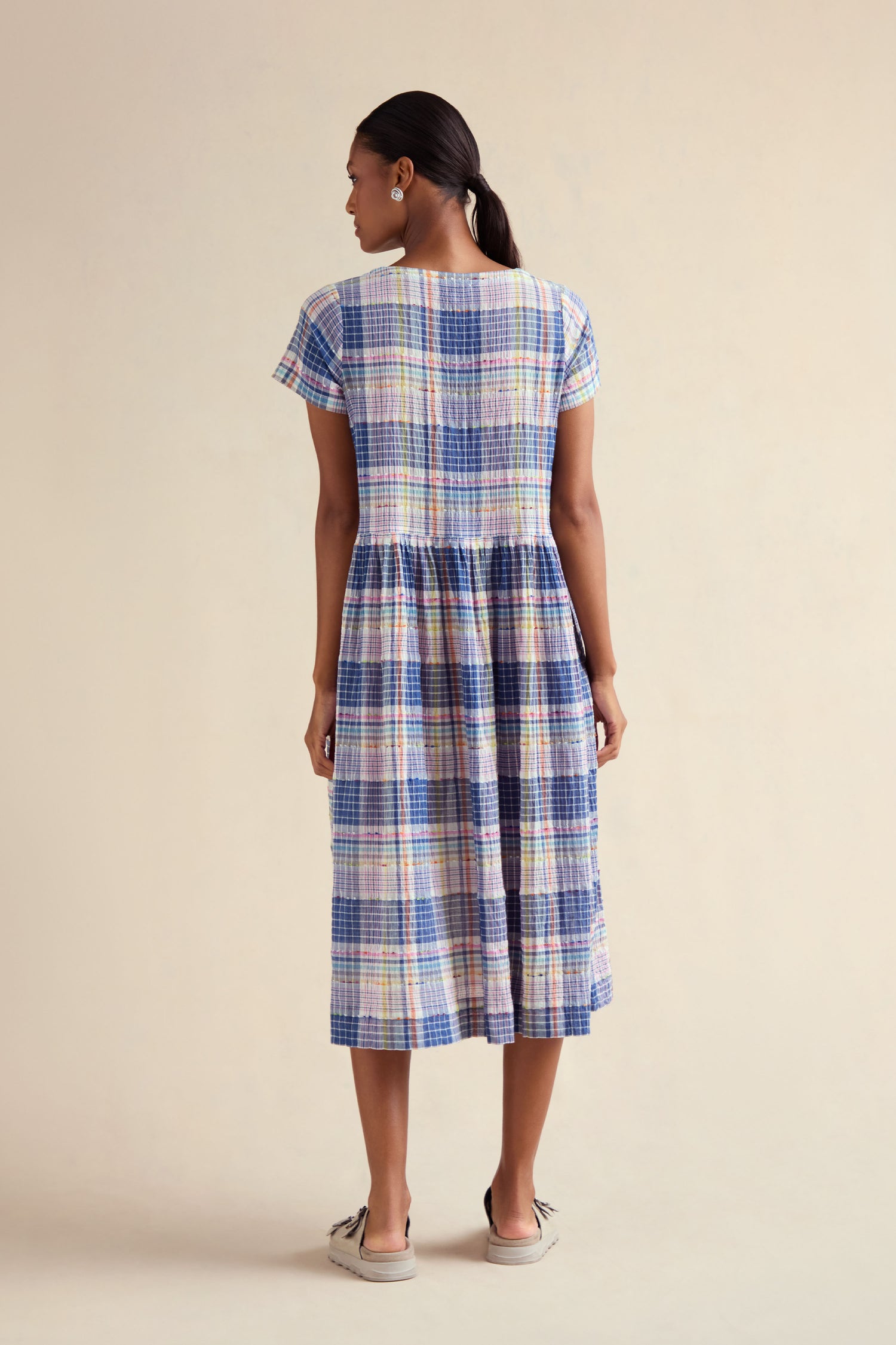 A woman stands with her back to the camera, wearing the Crinkle Check Jacquard Dress— a cotton-rich, short-sleeve A-line midi in pastel plaid—paired with casual shoes, set against a plain light background.