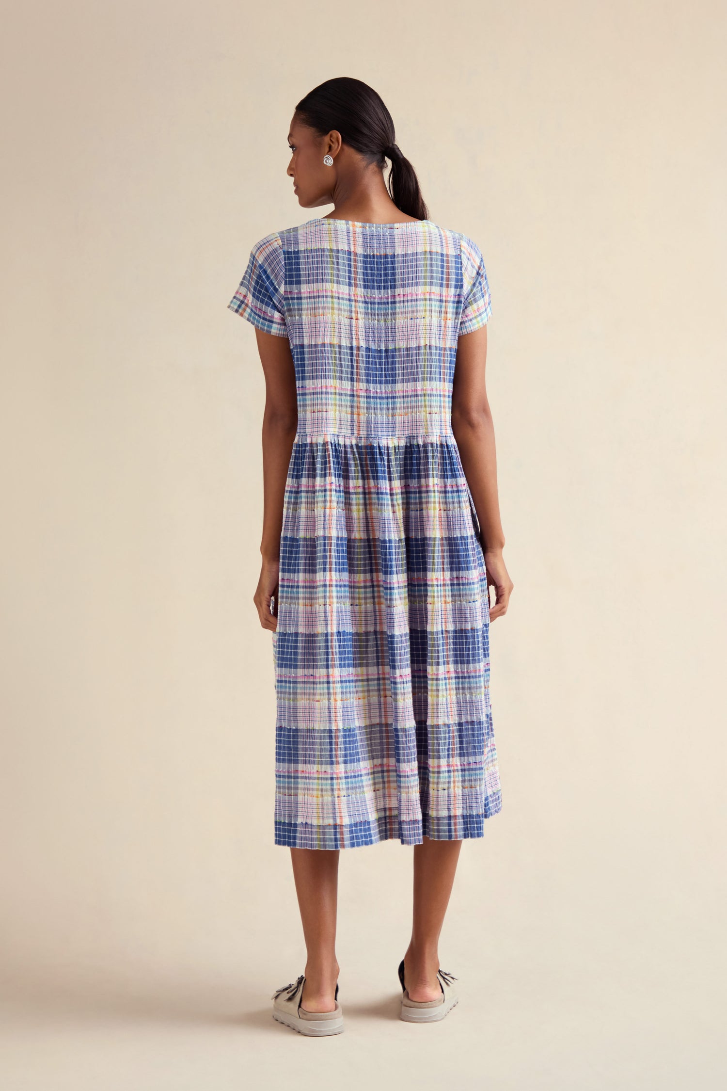 A woman with dark hair tied back, facing away, wears the Crinkle Check Jacquard Dress—short-sleeve, midi length with a gathered waist—and slip-on shoes.