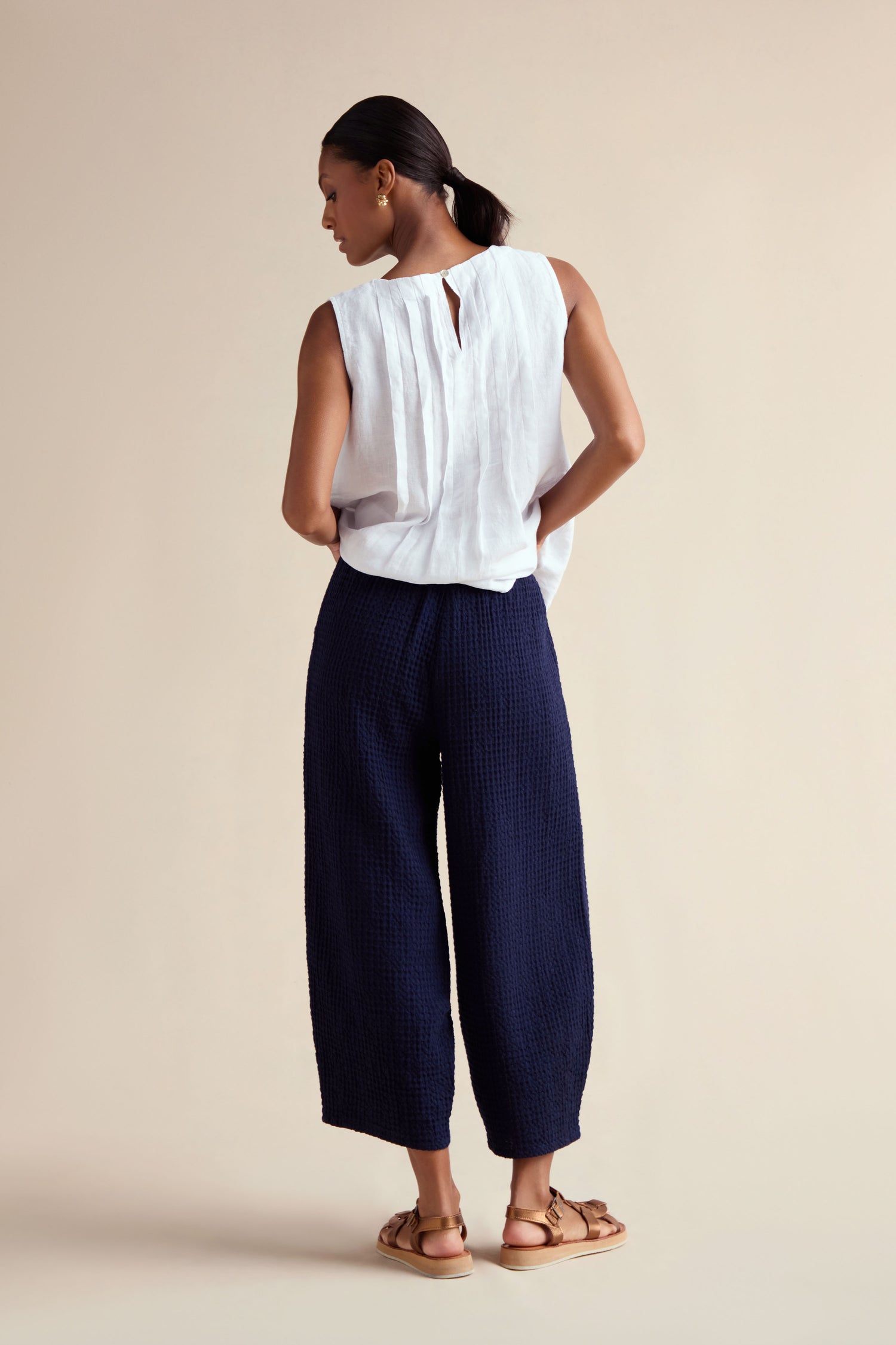 A woman stands with her back to the camera, wearing a sleeveless white top and cropped, dark blue Mini Waffle Cotton Bubble Trousers paired with sandals against a plain beige background.