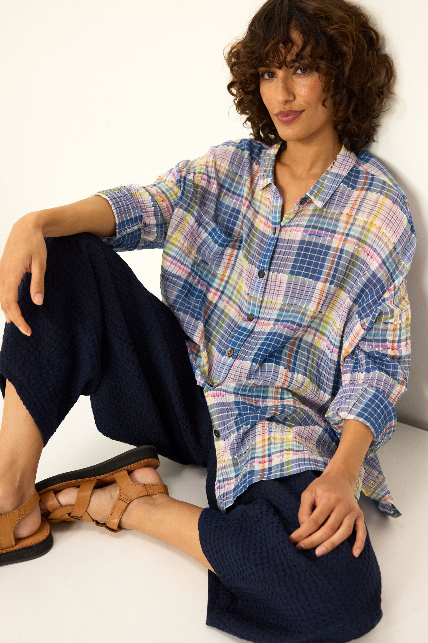 A person with curly brown hair sits against a white wall, wearing a loose plaid shirt, Mini Waffle Cotton Bubble Trousers in navy (cropped length), and tan sandals.