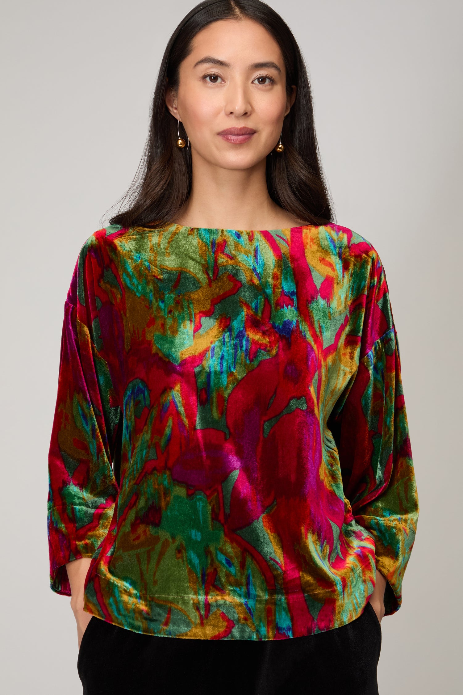 A woman with long dark hair wears gold earrings and the Velvet Blurred Botanicals Top, a versatile velvet top with red, green, pink, and yellow abstract patterns.