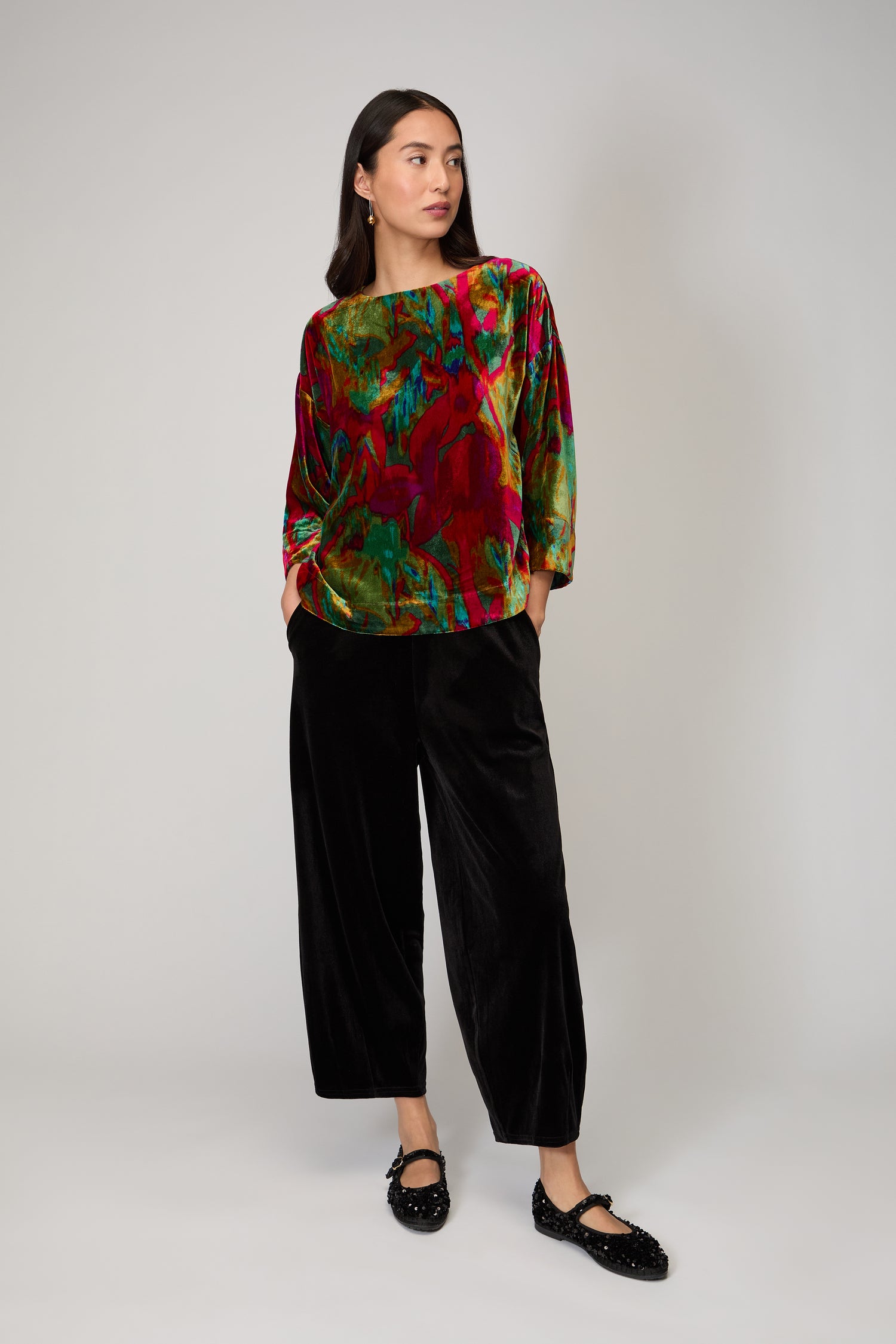 A woman stands against a plain background wearing the Velvet Blurred Botanicals Top, black velvet wide-leg pants, and black shoes.
