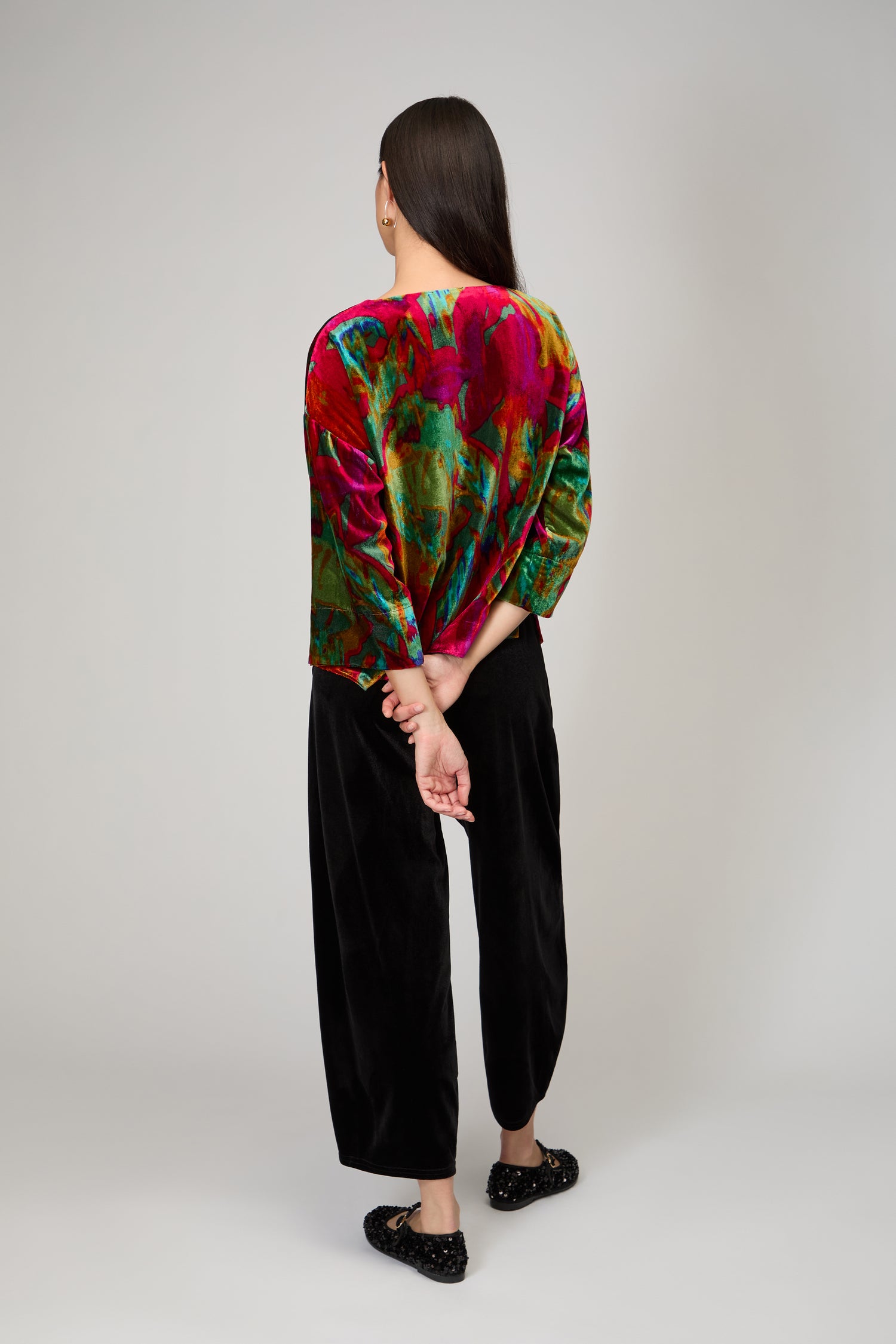 A person with long dark hair stands facing away against a plain background, wearing the Velvet Blurred Botanicals Top, paired with black pants and black shoes, hands clasped behind their back.