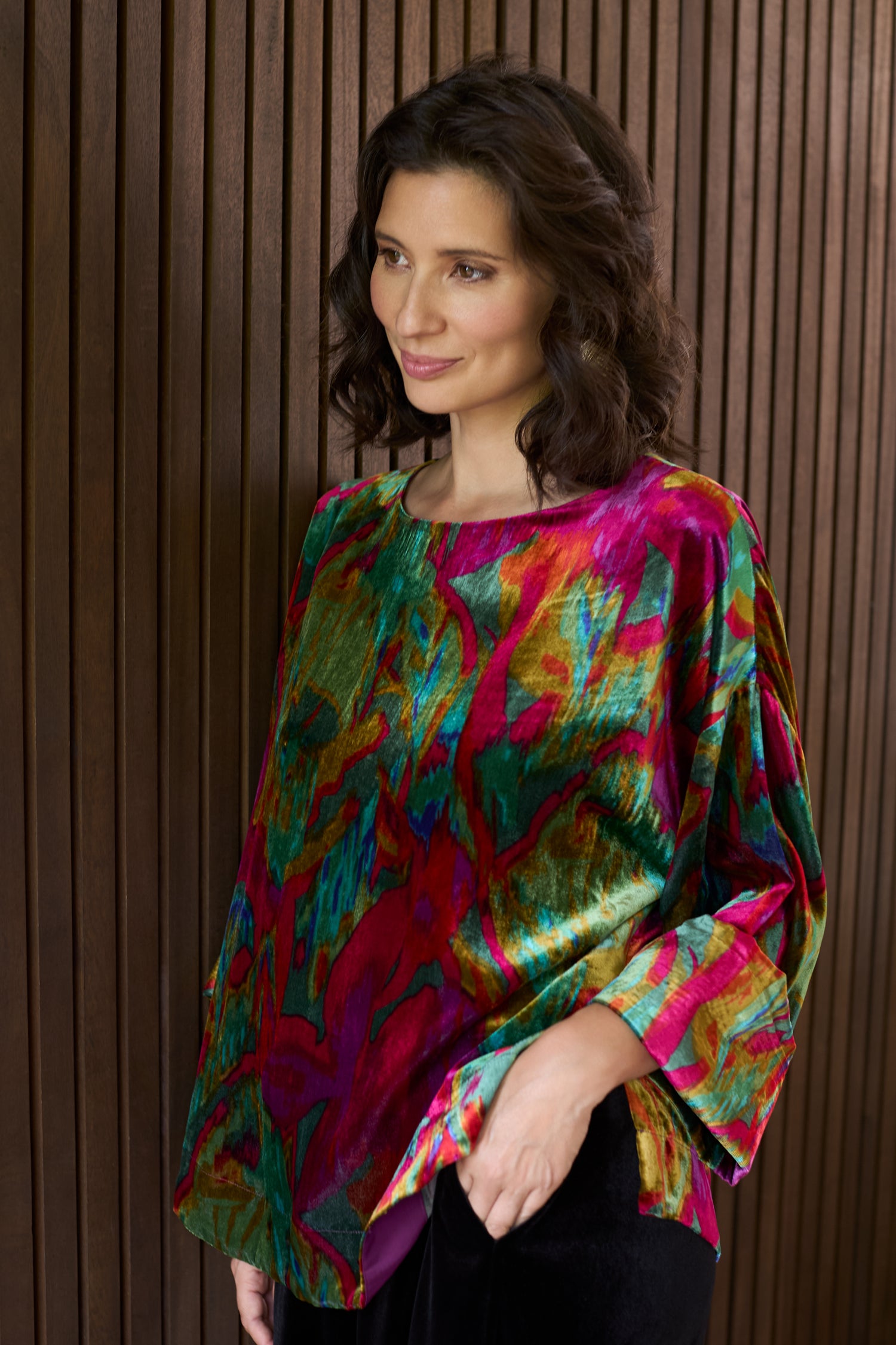 A woman with wavy dark hair stands before a wooden slat wall, wearing the Velvet Blurred Botanicals Top—a colorful, loose-fitting velvet piece featuring vibrant red, green, and yellow patterns.