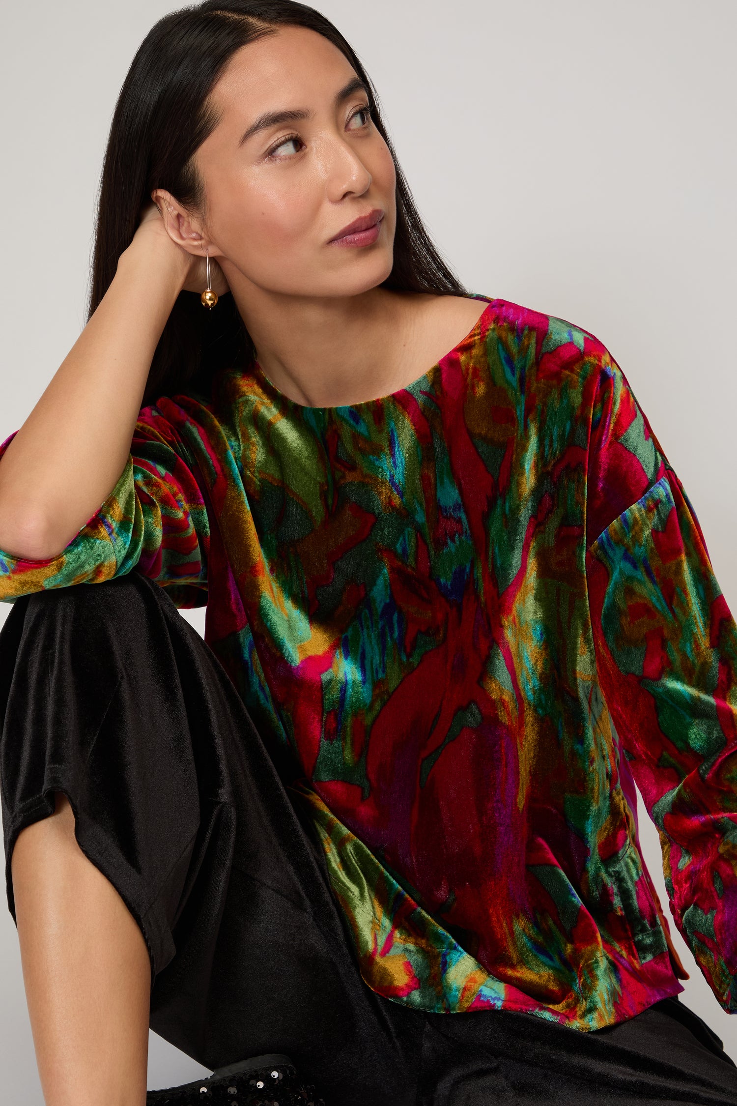 A woman with long dark hair wears the Velvet Blurred Botanicals Top and black pants, sitting with one arm on her knee, gazing thoughtfully to the side.