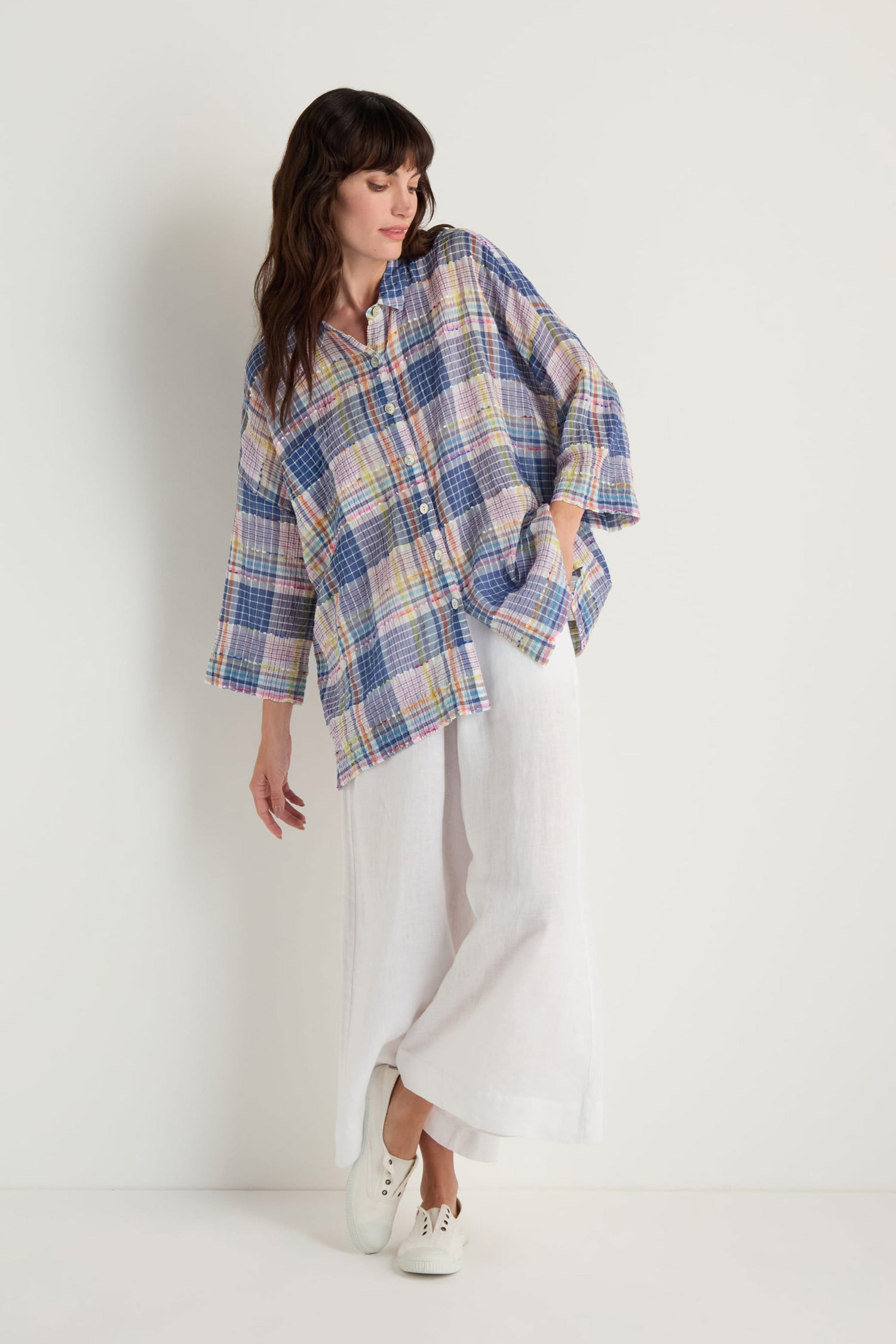A woman stands against a plain white background, looking slightly to the side, wearing a loose, boxy fit Crinkle Check Jacquard Shirt in multicolored checks with wide-legged white pants.