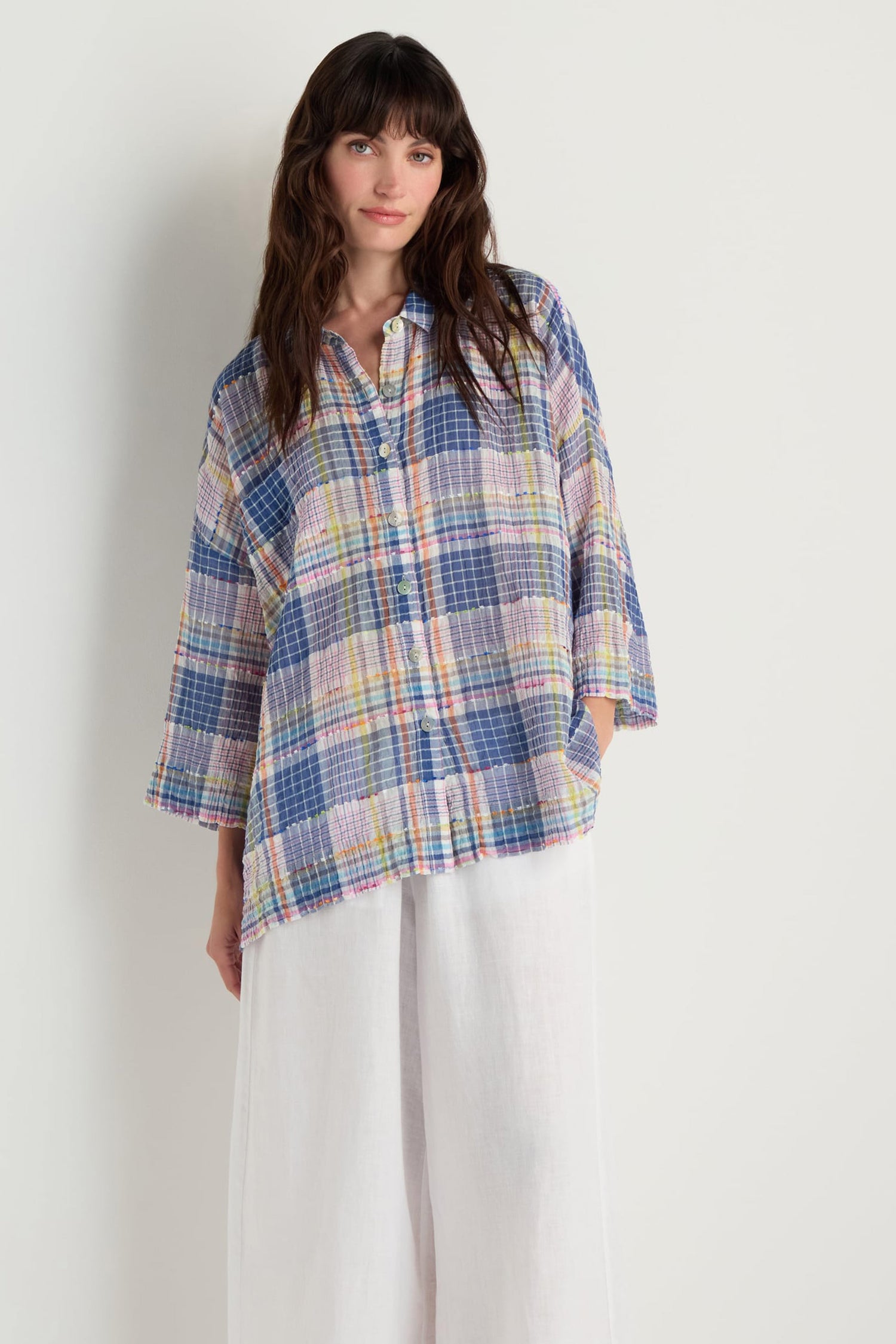 A woman stands against a plain background wearing the Crinkle Check Jacquard Shirt in a boxy fit with pastel checks, paired with wide-leg white pants.