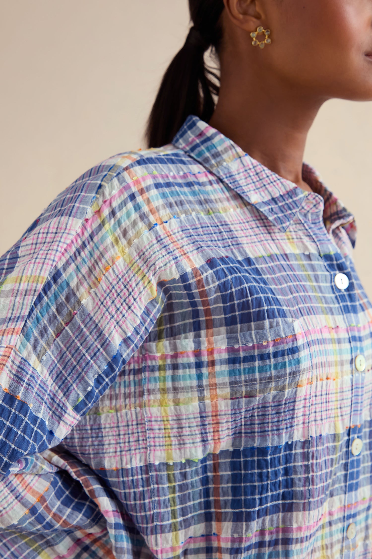 A close-up of a person with dark hair tied back and wearing a flower-shaped earring, dressed in the Crinkle Check Jacquard Shirt featuring a multicolored pattern and white buttons.