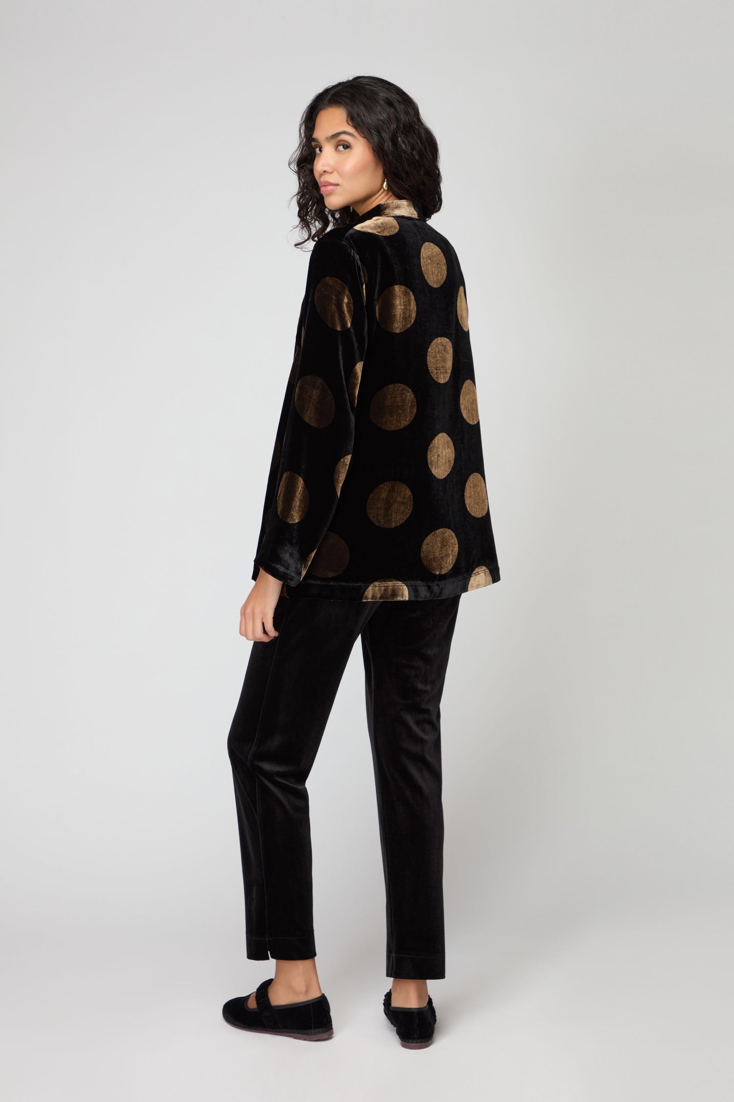 A person with curly hair faces away, wearing the Silk Velvet Spot Shirt in black with gold spots and matching loafers, against a light gray background. This chic statement top adds flair to their stylish look.