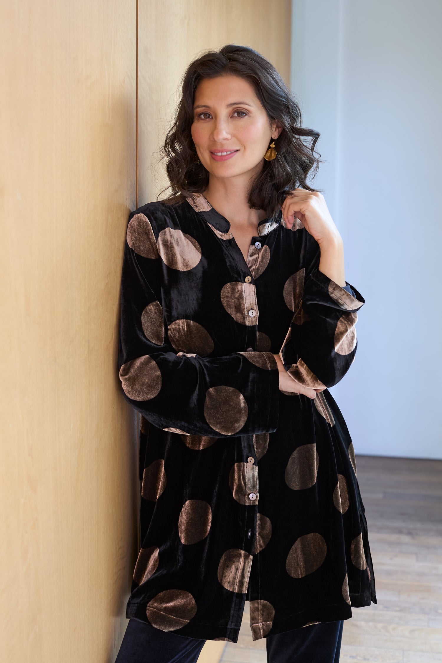A woman with wavy brown hair stands indoors, leaning against a light wood wall, wearing the Silk Velvet Spot Shirt—a versatile black velvet top with large beige polka dots.