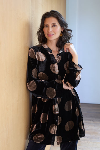 A woman with wavy brown hair stands indoors, leaning against a light wood wall, wearing the Silk Velvet Spot Shirt—a versatile black velvet top with large beige polka dots.