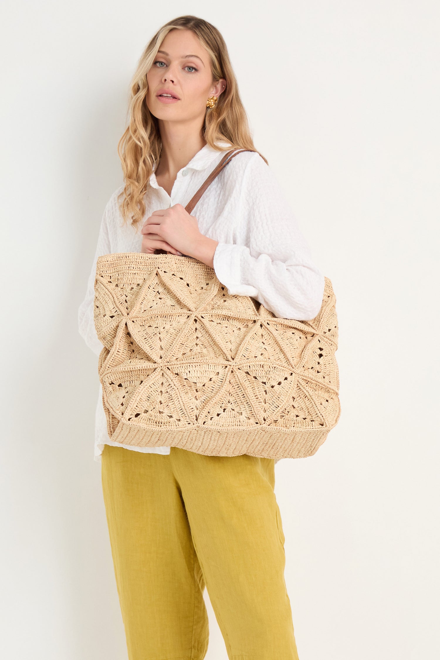 A woman in a white blouse and yellow pants holds the Handwoven Raffia Basket Bag—a large, textured beige tote with a geometric pattern—over her shoulder.