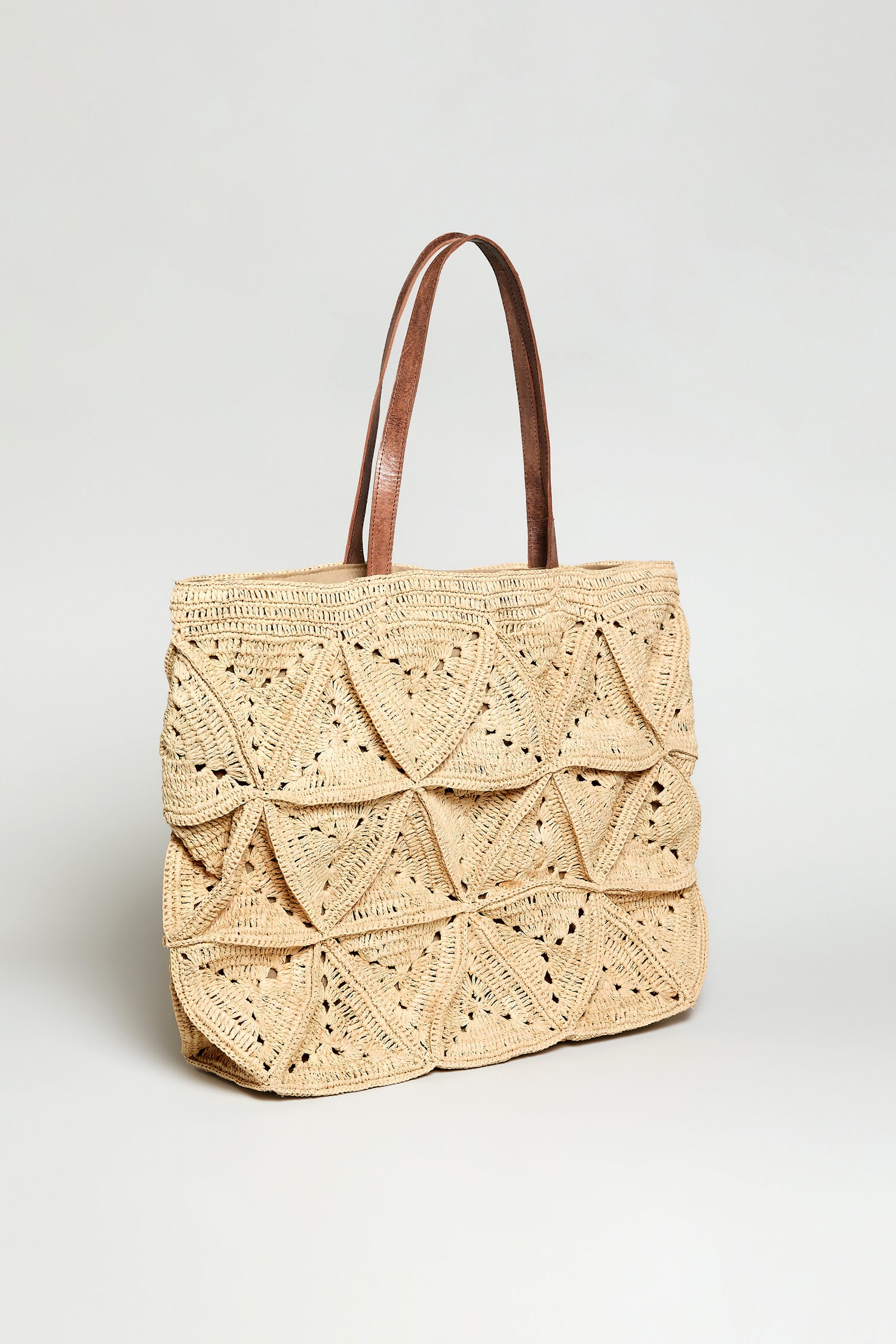 The Handwoven Raffia Basket Bag, with brown leather handles and a geometric-patterned body, is shown against a plain light background.