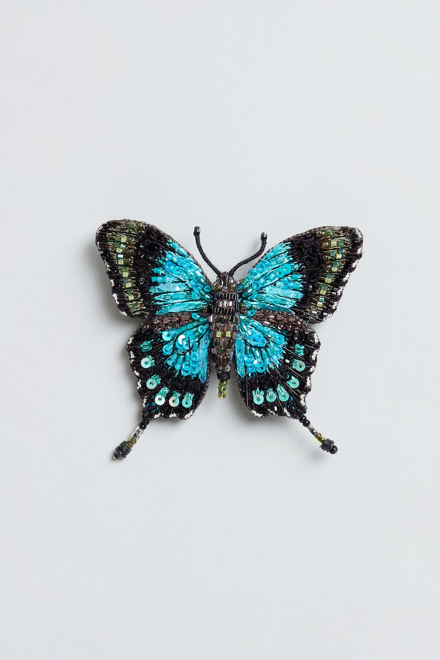 A hand beaded sea green swallowtail brooch, meticulously adorned with beads and sequins, features striking turquoise and black wings against a light grey background.