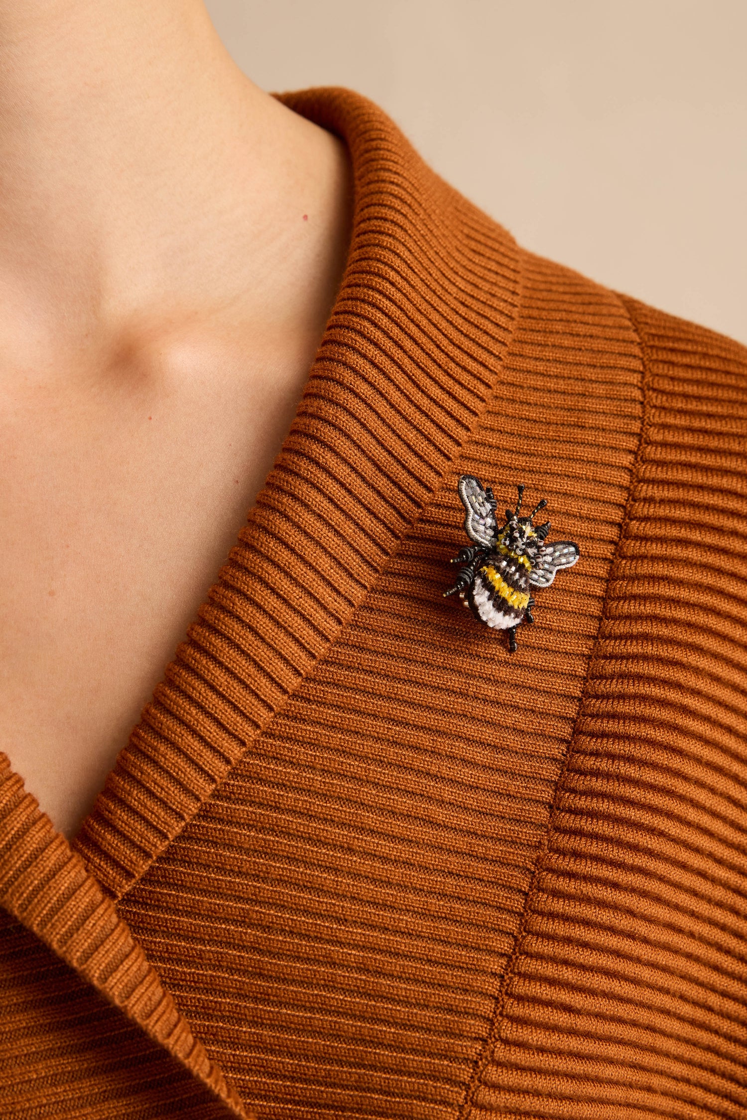 A person wearing a ribbed, brown sweater with a Hand Beaded Yellow-Banded Bee Brooch pinned to the left side near the collarbone.