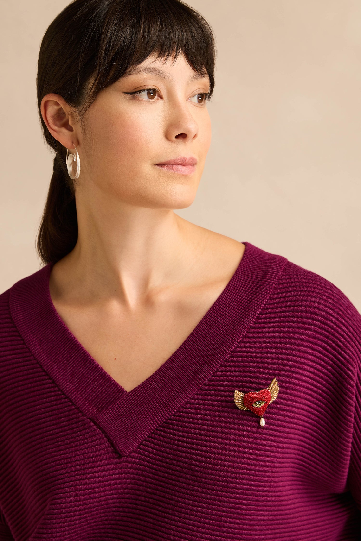A model with dark hair in a ponytail wears silver hoop earrings and a ribbed purple v-neck sweater, accented with a stunning Hand Beaded Guardian Heart Brooch—an intricately hand beaded accessory featuring delicate wings and a central eye detail.