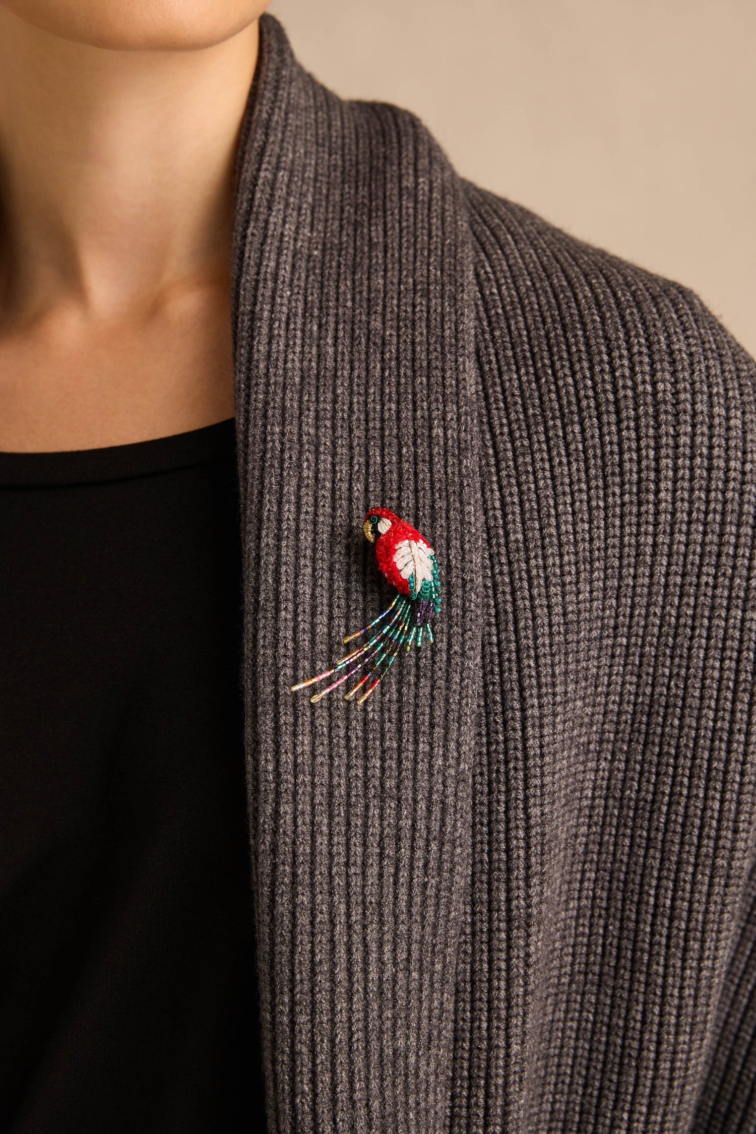 The Hand Beaded Arara Macao Brooch, inspired by the vibrant scarlet macaw and featuring a multicolored beaded tail, is pinned to the lapel of a grey ribbed knit cardigan.