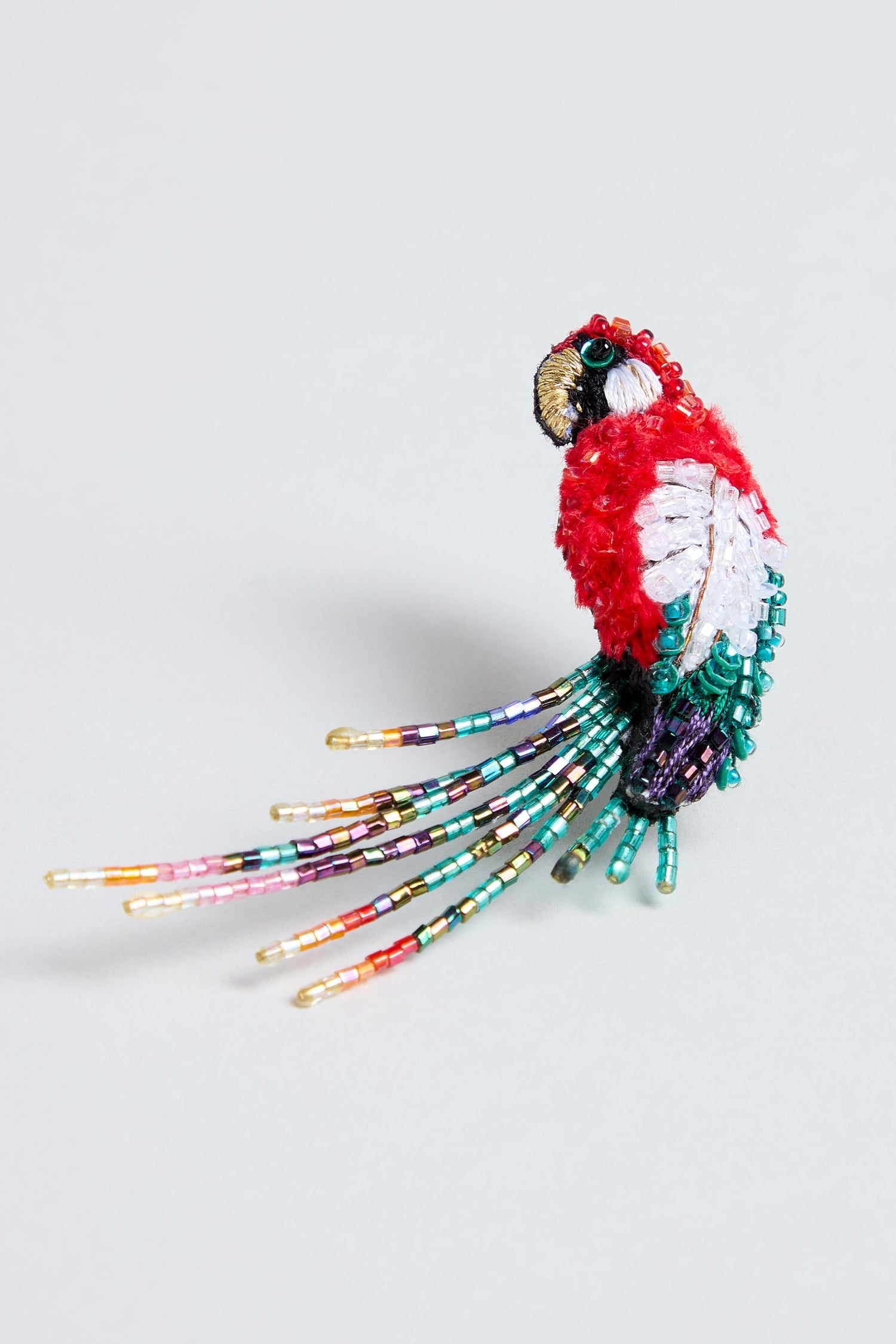 Introducing the Hand Beaded Arara Macao Brooch: a stunning accessory shaped like a parrot and adorned with red, white, green, purple, and gold beads. This brooch features long, colorful beaded tail feathers set against a light background.