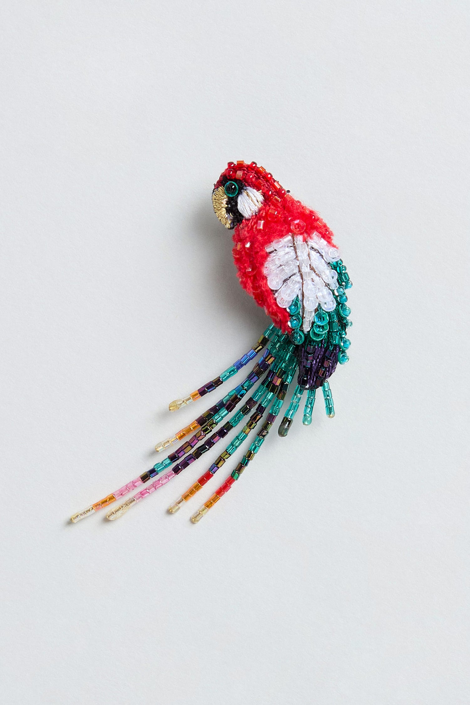 Discover the Hand Beaded Arara Macao Brooch, a beautifully crafted piece featuring red, white, and teal beads accented by long, dangling beaded tail feathers—all exquisitely set against a light gray background.