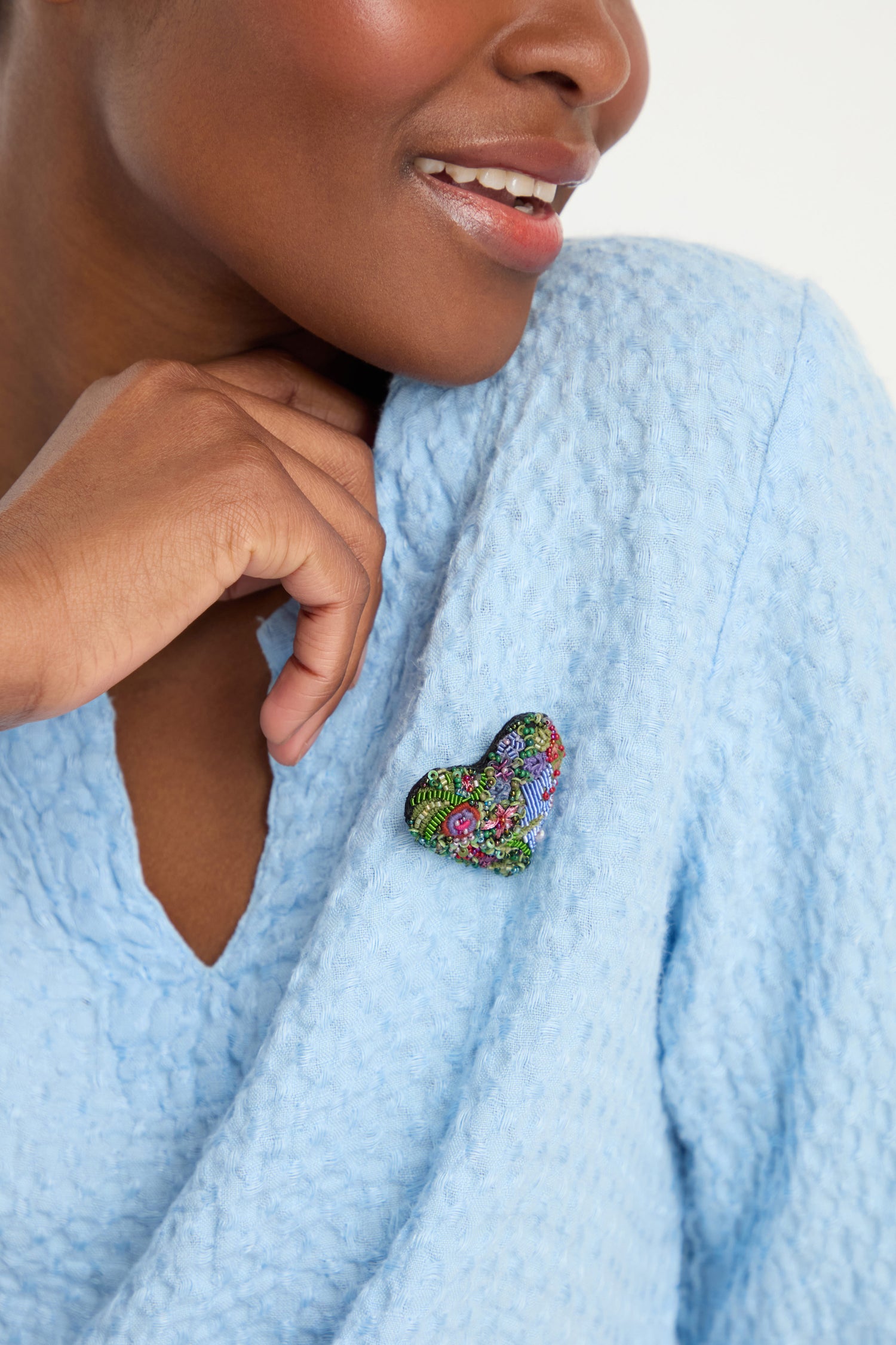 A person in a textured light blue top wears the Hand Embroidered Blooming Heart Brooch, full of colorful details, pinned near their shoulder.