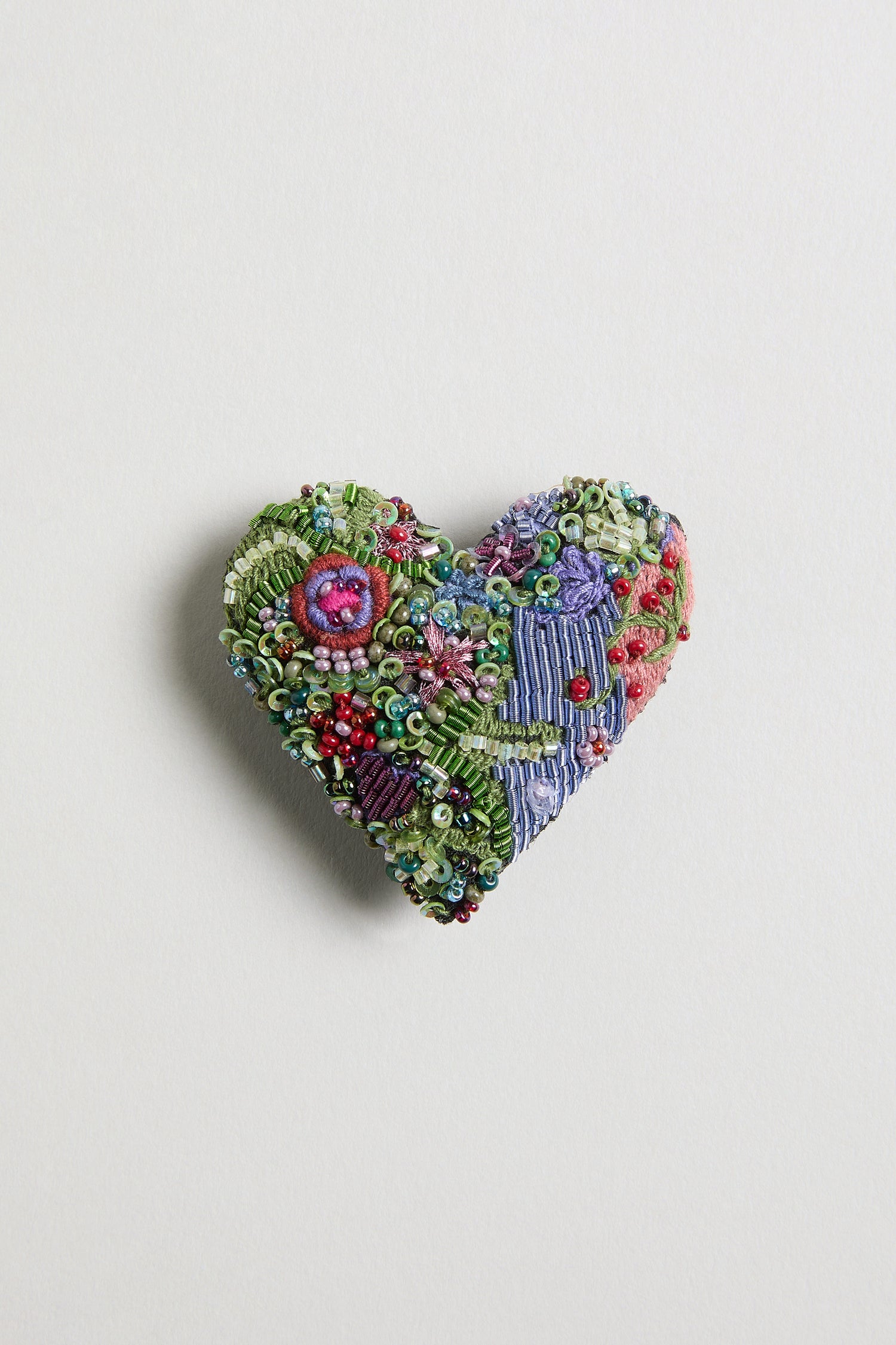 The Hand Embroidered Blooming Heart Brooch features a heart shape with vibrant, detailed embroidery and beadwork on a neutral background.