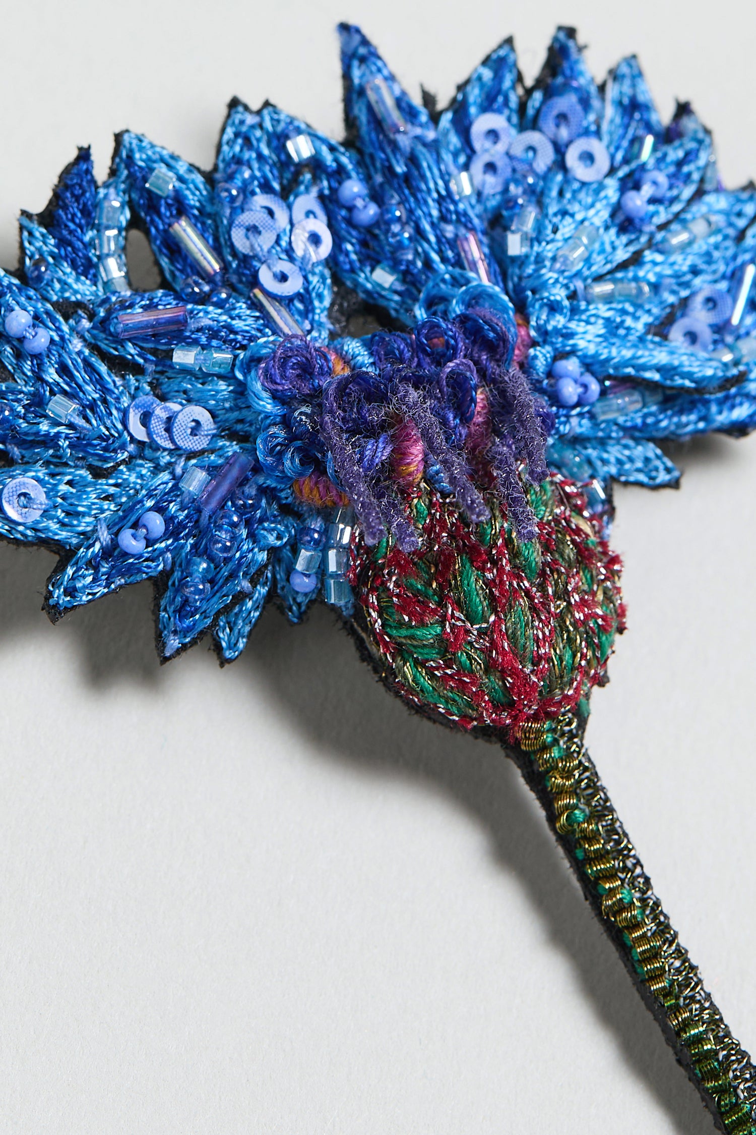 Close-up of the Hand Embroidered Blue Cornflower Brooch, adorned with sequins and beads and featuring a green and black stem—a stunning piece of botanical jewelry displayed on a light background.