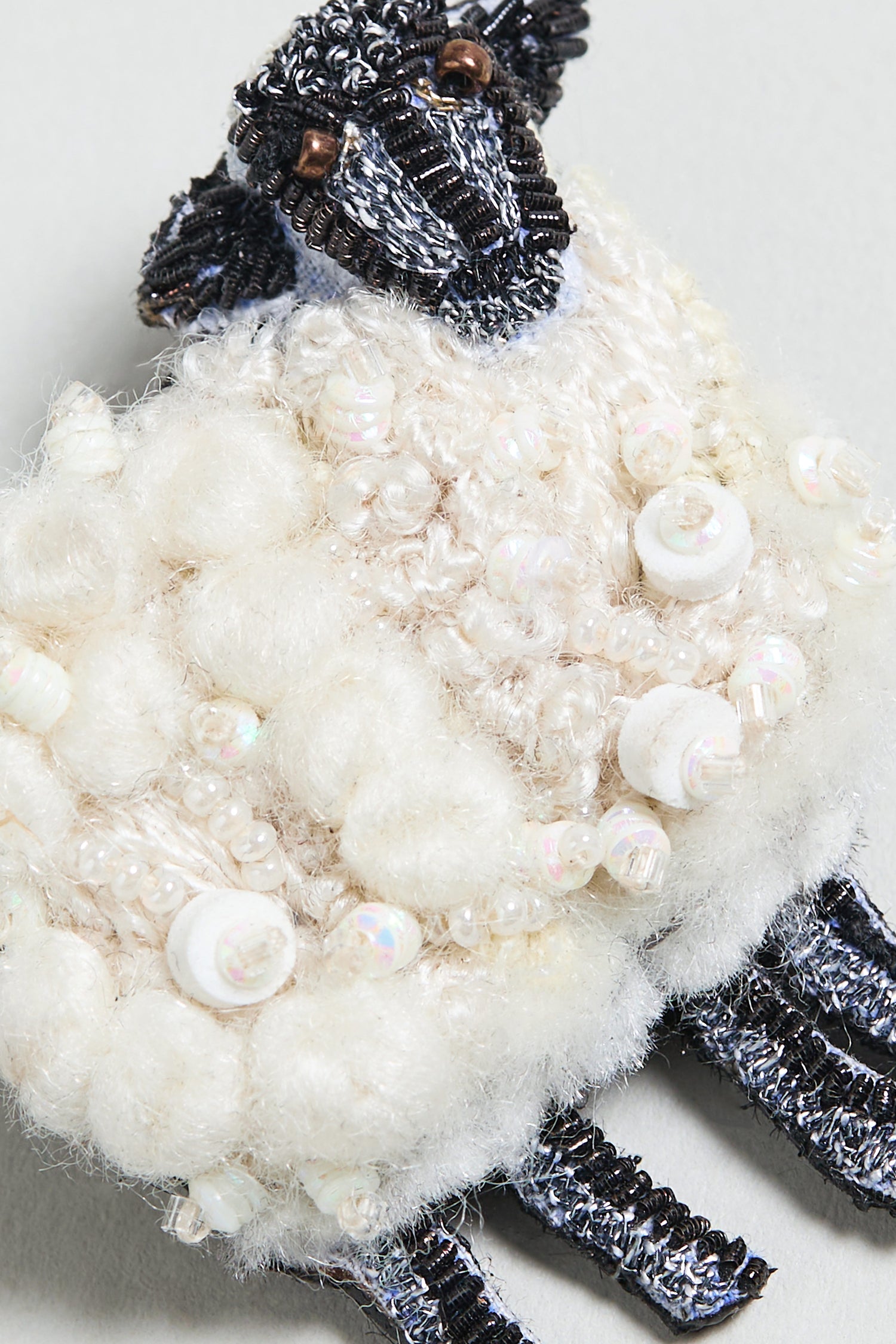 The Hand Embroidered Sheep Brooch features a collectible sheep design, crafted with white wool, beads, and sequins for texture, and detailed with black beads on the head and legs.