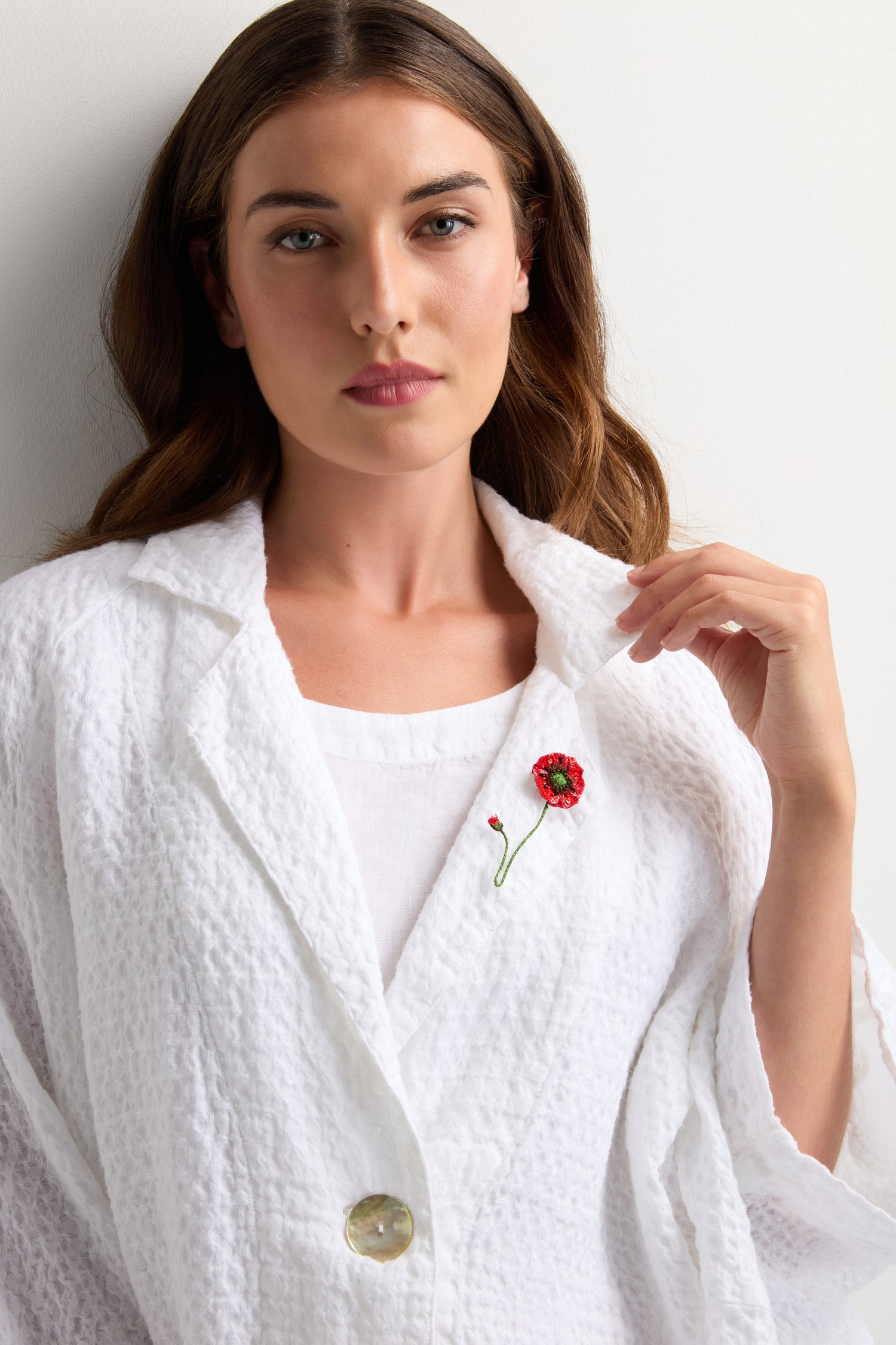 A woman in a textured white blazer and top wears the Hand Beaded Shirley Poppy Brooch on her lapel, standing against a plain light background.