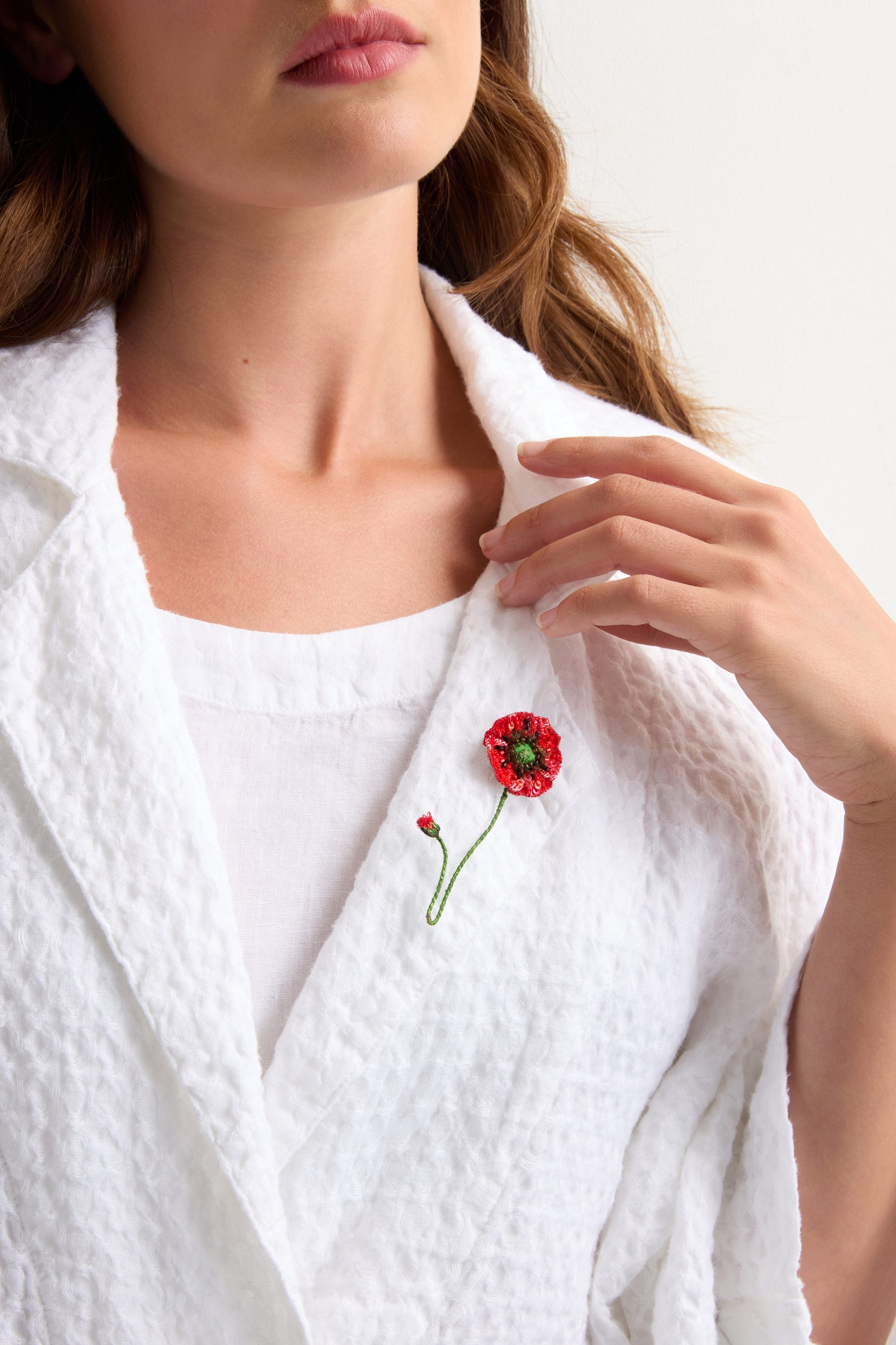 A woman in a textured white outfit with botanical flair—a Hand Beaded Shirley Poppy Brooch is pinned to her lapel.