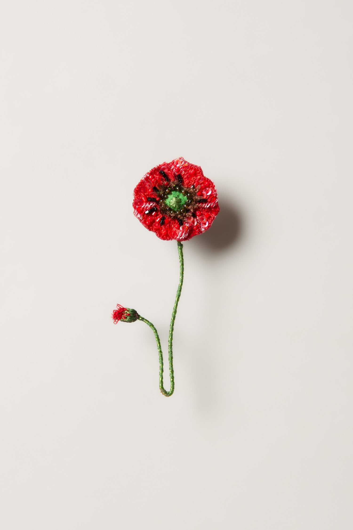The Hand Beaded Shirley Poppy Brooch features a red flower with a green stem and small bud on a light background—perfect for lovers of handcrafted accessories and botanical jewelry.