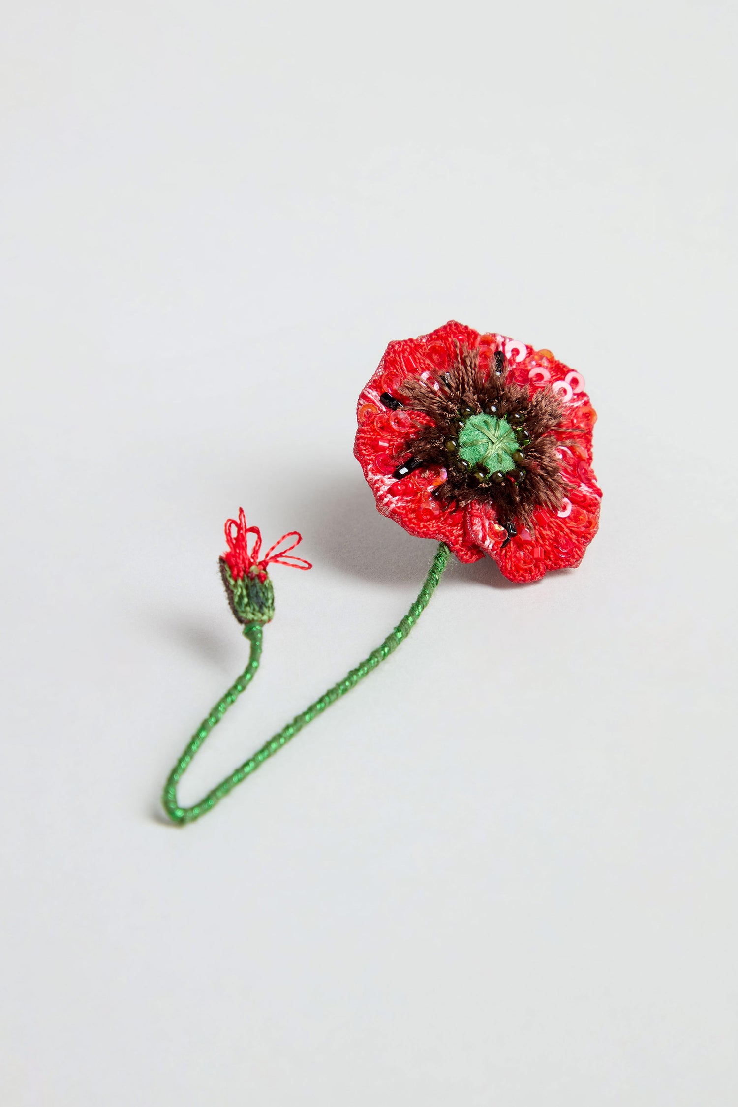 The Hand Beaded Shirley Poppy Brooch, featuring a vibrant red flower and green stem made from beads and sequins, rests on a plain light background.