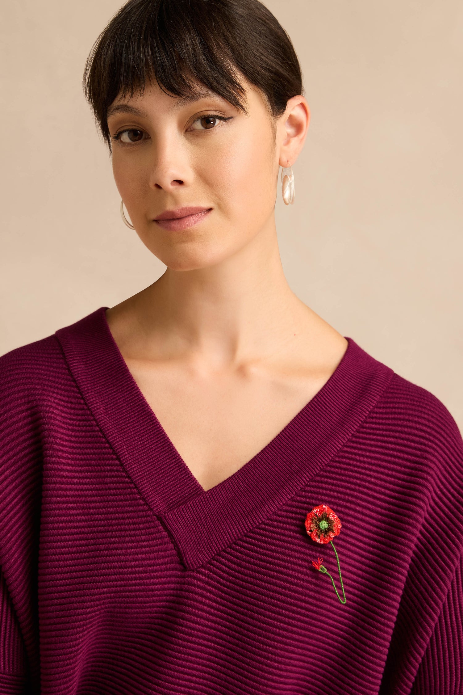 A woman with short brown hair wears a deep purple V-neck sweater and hoop earrings, with a Hand Beaded Shirley Poppy Brooch pinned to her sweater.