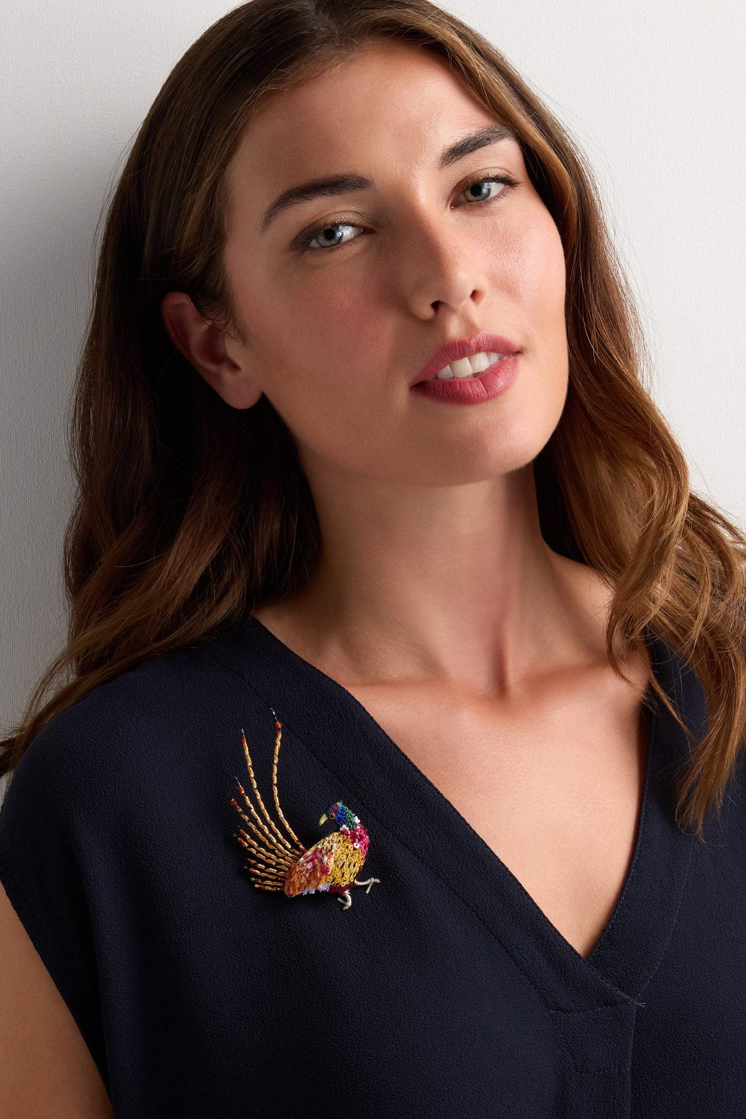 A woman with long brown hair wears a navy V-neck top adorned with the Hand Beaded Pheasant Brooch on her left side—a colorful accessory, perfect as a gift for nature lovers.