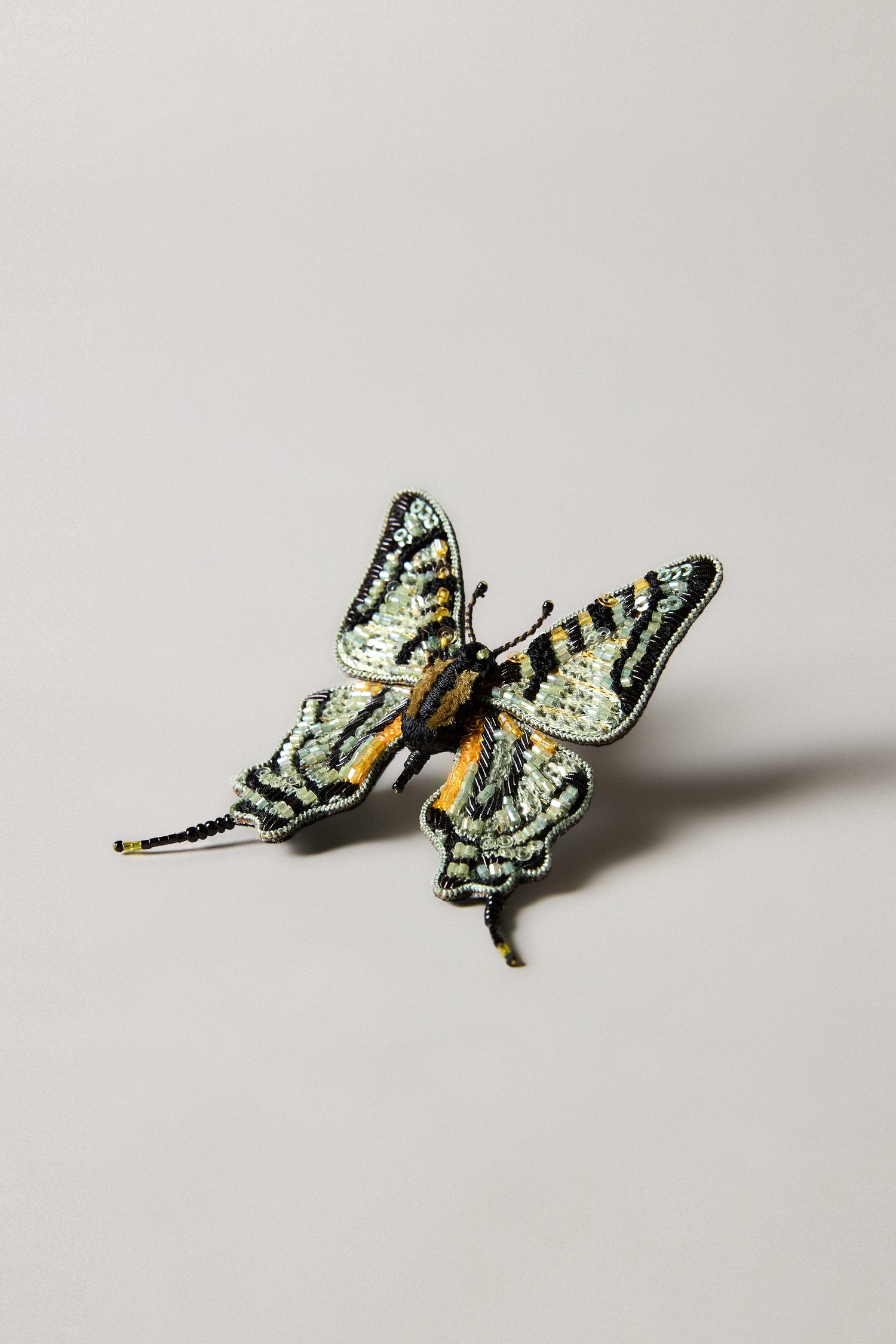 A Hand Beaded Swallowtail Brooch from Trovelore, featuring intricate black, yellow, and green embroidery, is displayed on a neutral background.