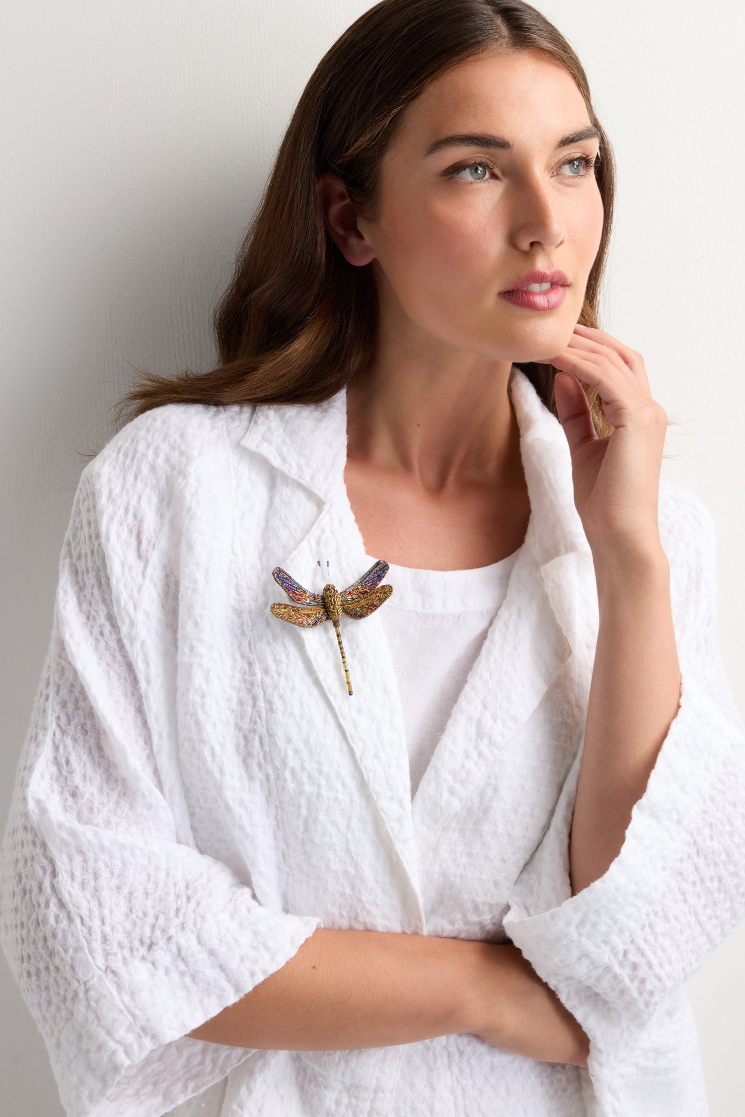 A woman in a white textured, hand-stitched garment wears the Hand Beaded Hawker Dragonfly Brooch—an exquisite piece of wearable art—while gazing to the side with her hand near her chin.