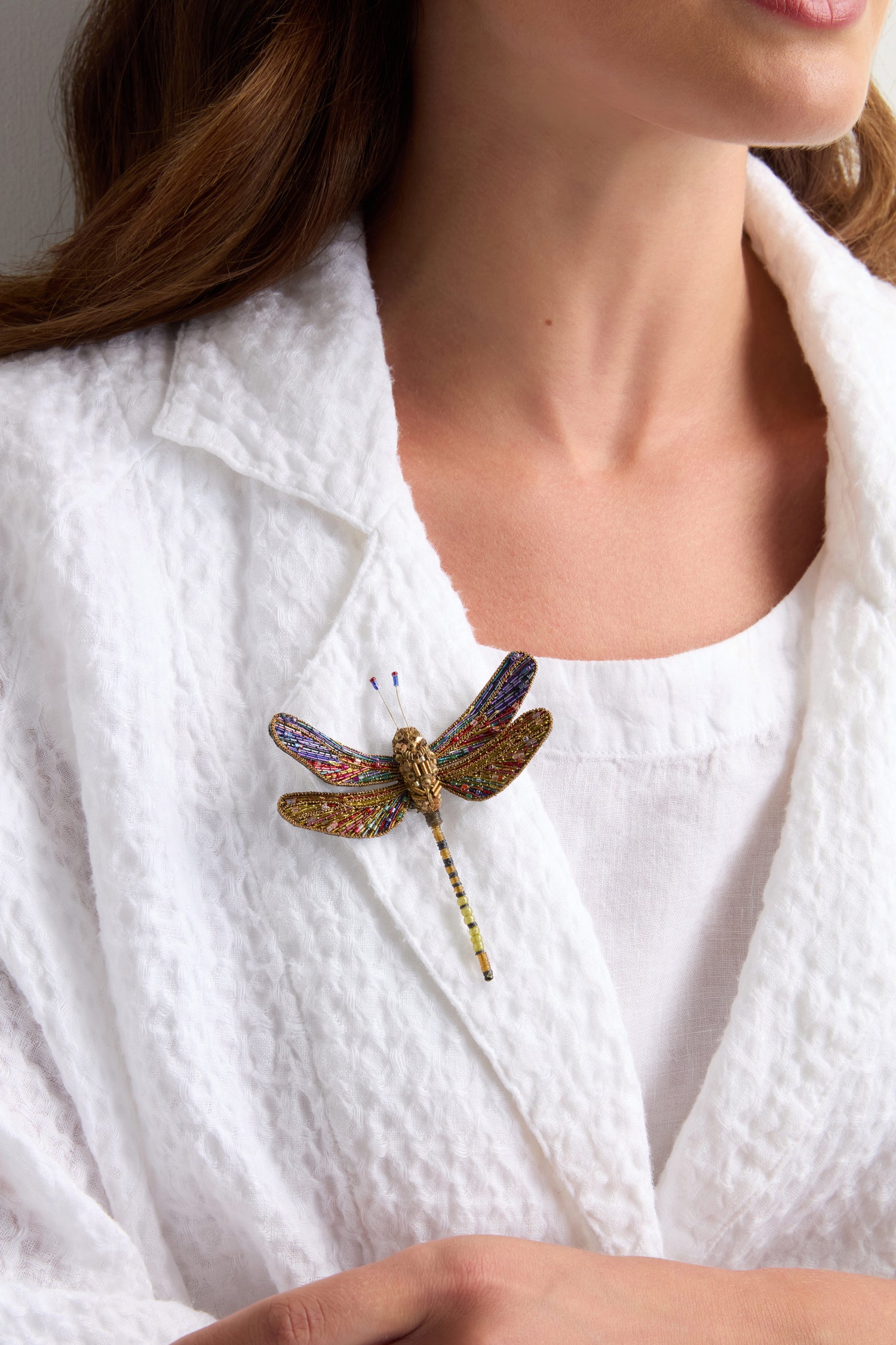 A woman wears a white textured blazer and top, with the Hand Beaded Hawker Dragonfly Brooch pinned to her lapel.