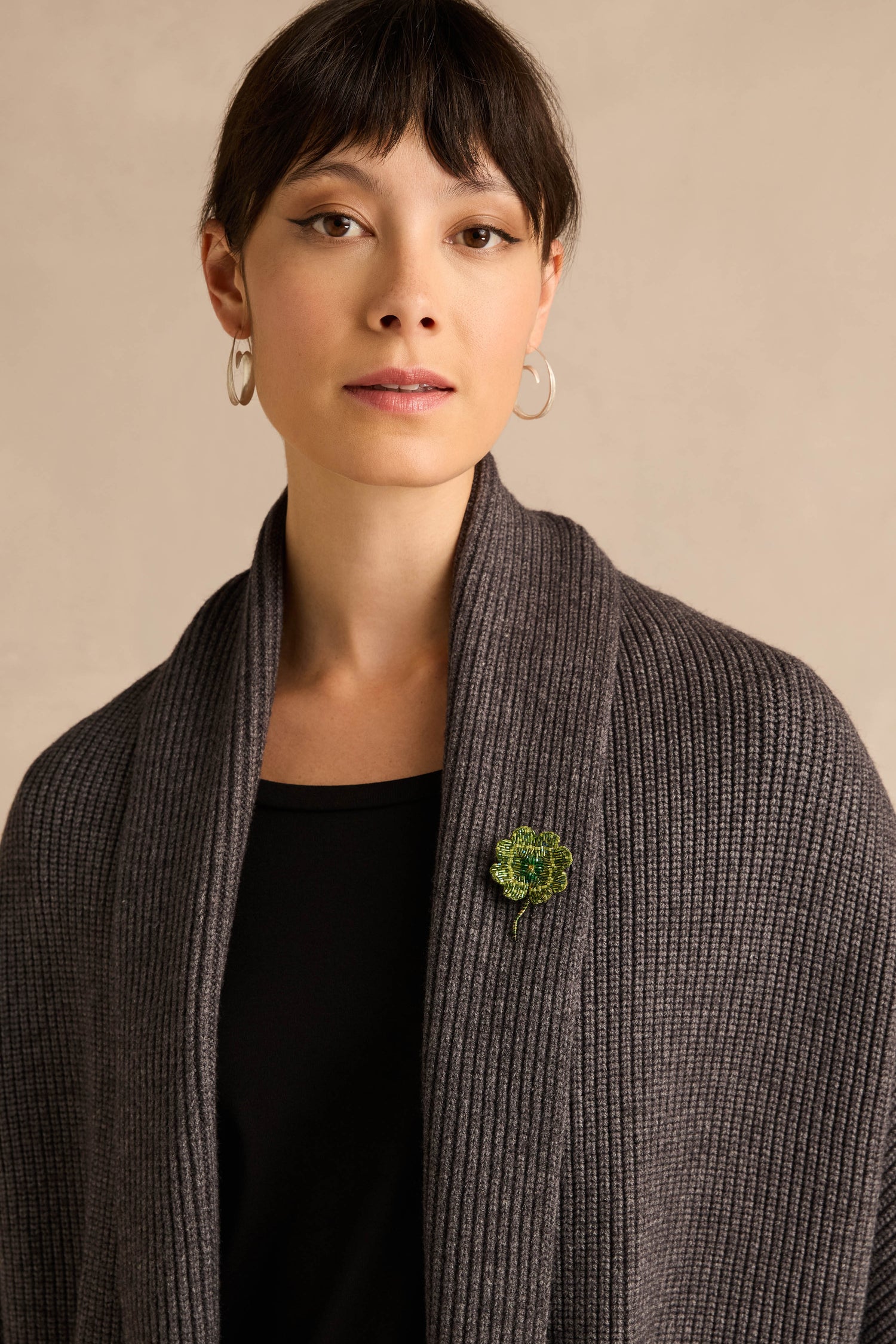 A woman with short brown hair, silver hoop earrings, a ribbed gray shawl, and a black top stands against a neutral background, her shawl adorned with the Hand Beaded Vintage Clover Brooch.