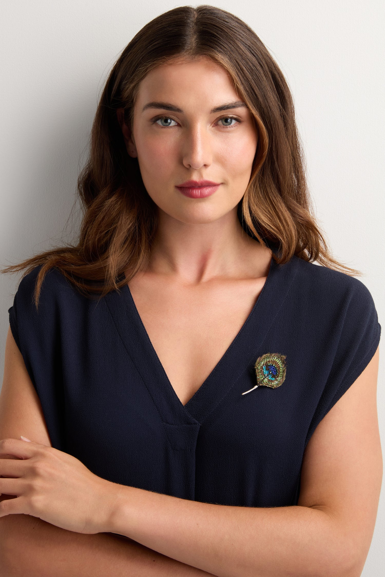 A woman with long brown hair wears a navy V-neck top and the Hand Beaded Peacock Feather Brooch on her left side, standing against a plain light background.