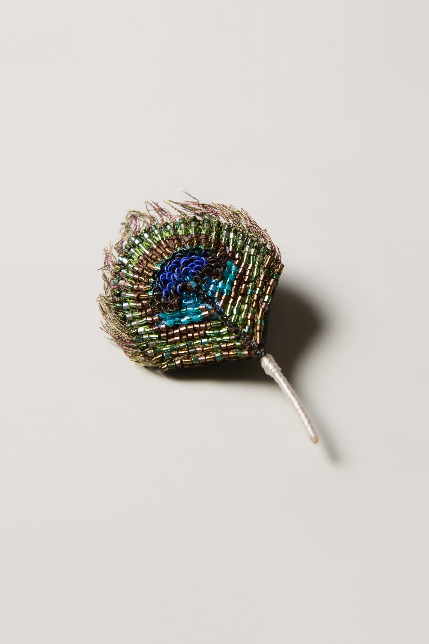 The Hand Beaded Peacock Feather Brooch features green, blue, and gold beads with a short white stem, shown on a plain light background.